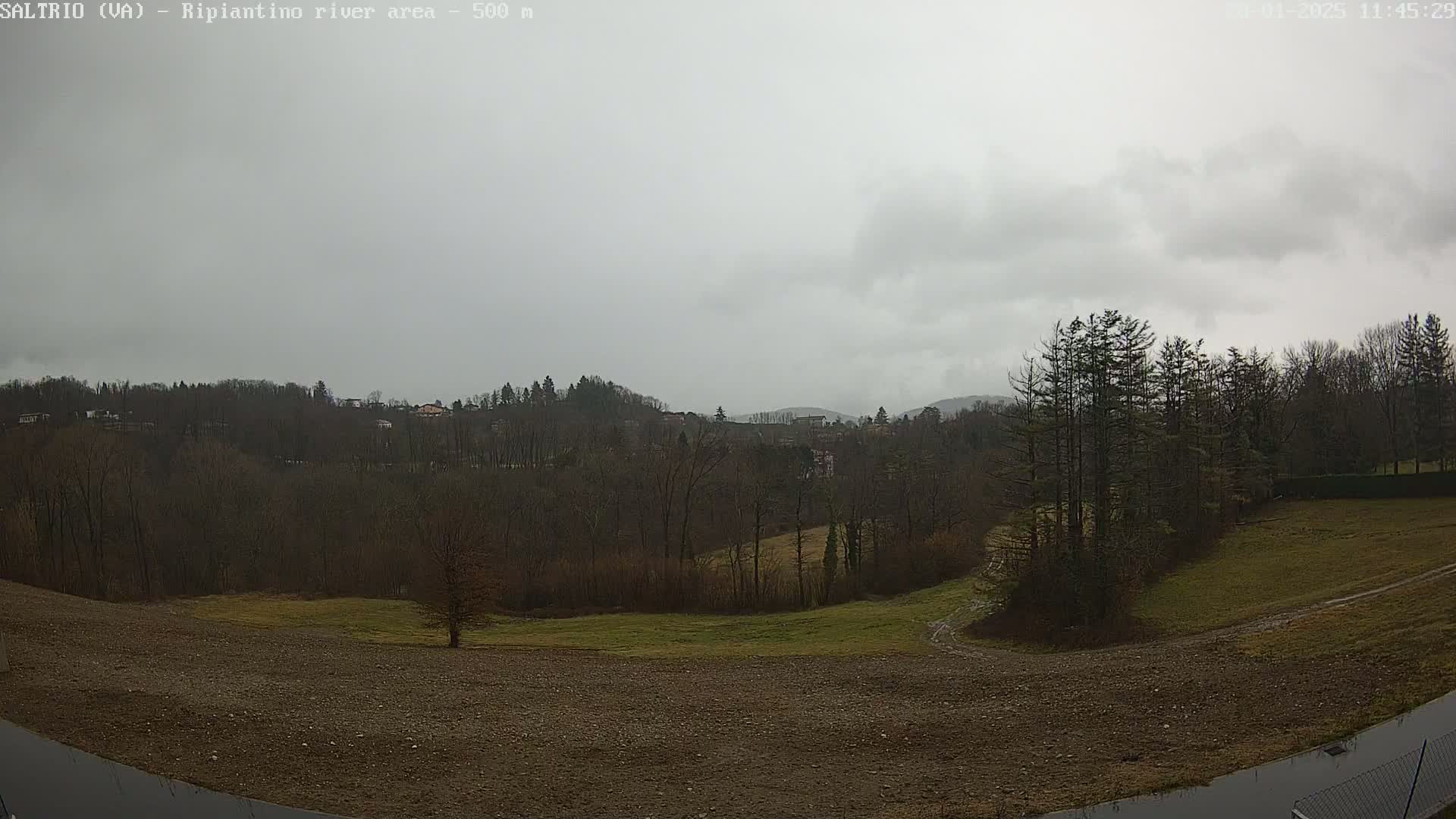 A view of a hilly landscape under an overcast sky shows bare trees and a field of brown earth in the foreground.