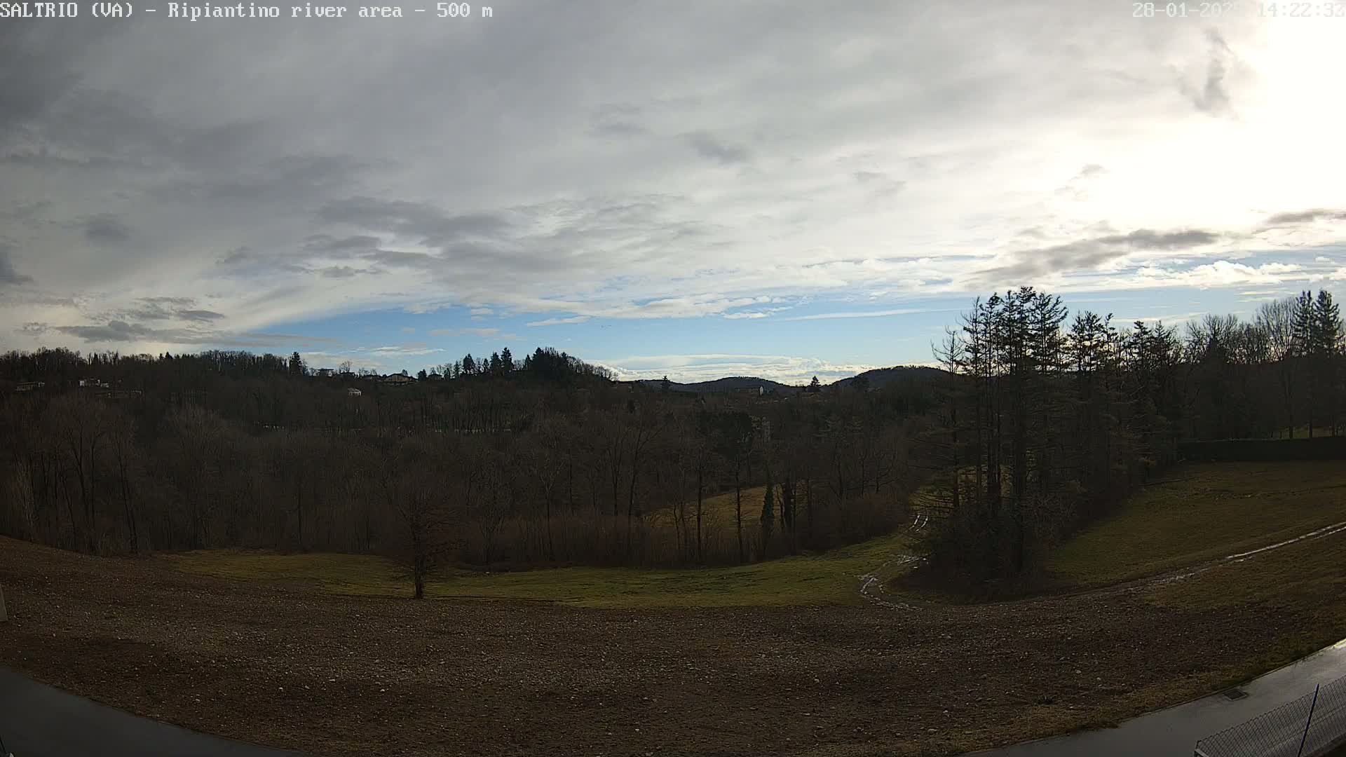 A partly cloudy sky overlooks a mostly barren, hilly landscape with dark-colored trees and patches of brown and green fields.