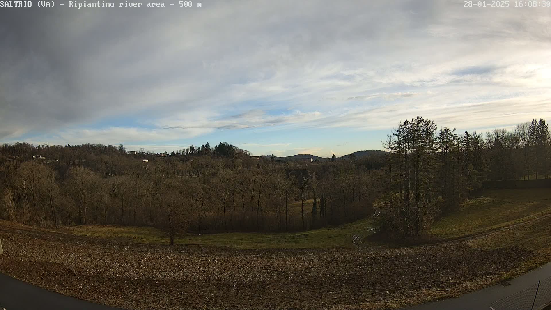 A mostly cloudy sky overlooks a sparsely wooded landscape with a field in the foreground.