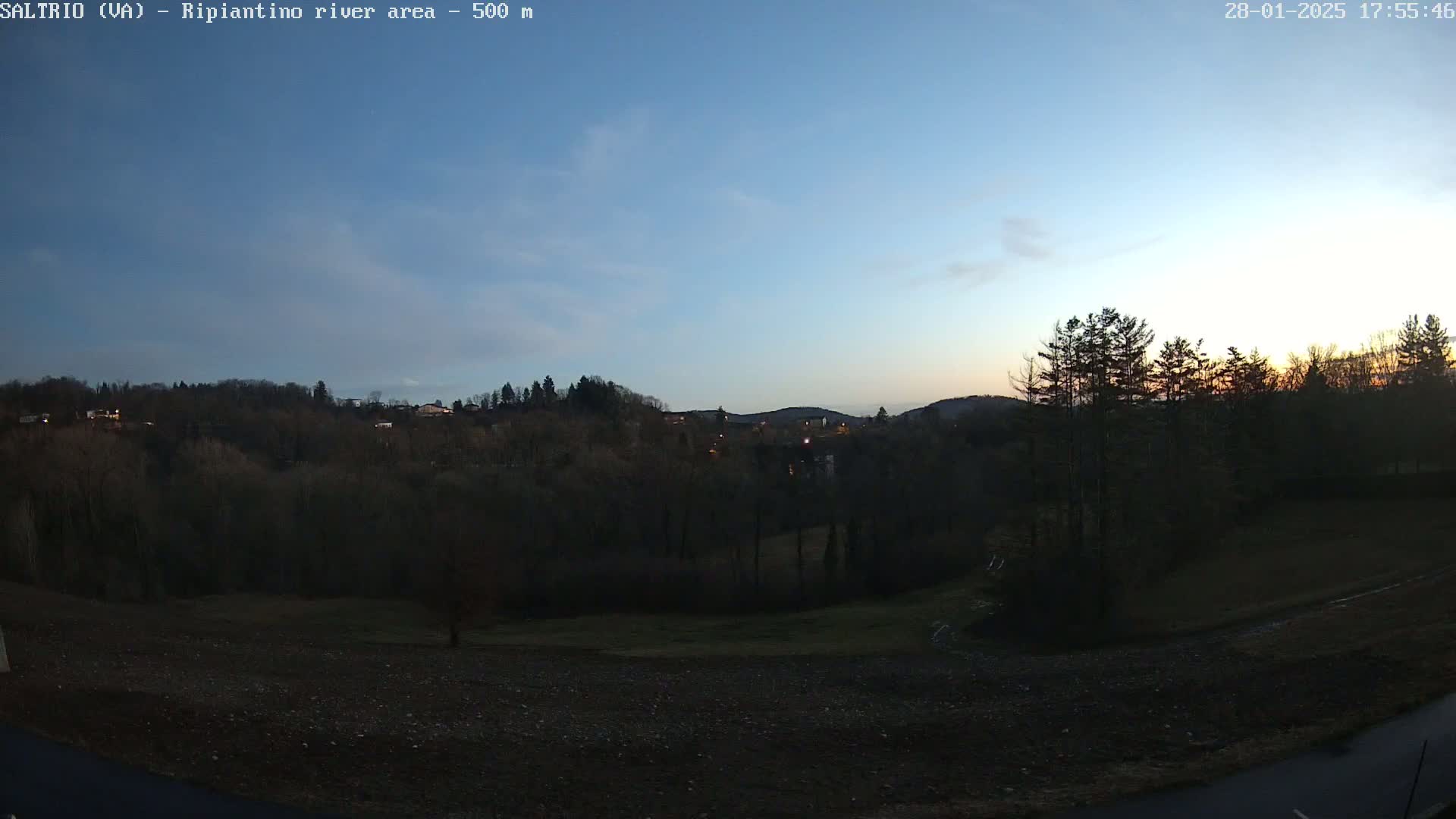 A mostly clear twilight sky over a dark, wooded hillside with a few scattered lights visible in the distance.
