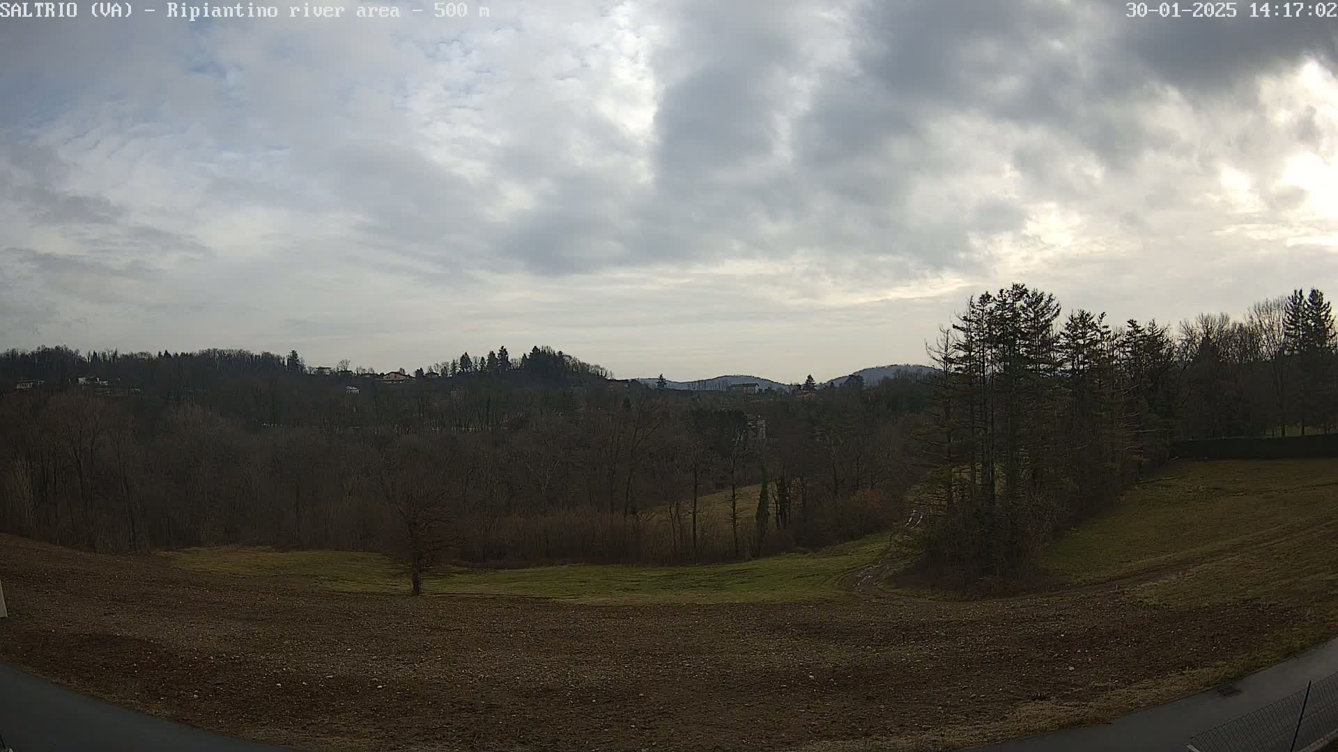 A mostly cloudy day reveals a view of a plowed field, bare winter trees, and distant hills.