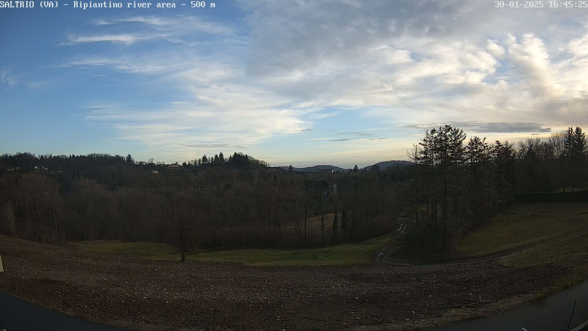 A partly cloudy sky overlooks a hilly landscape with bare trees and a plowed field in the foreground.