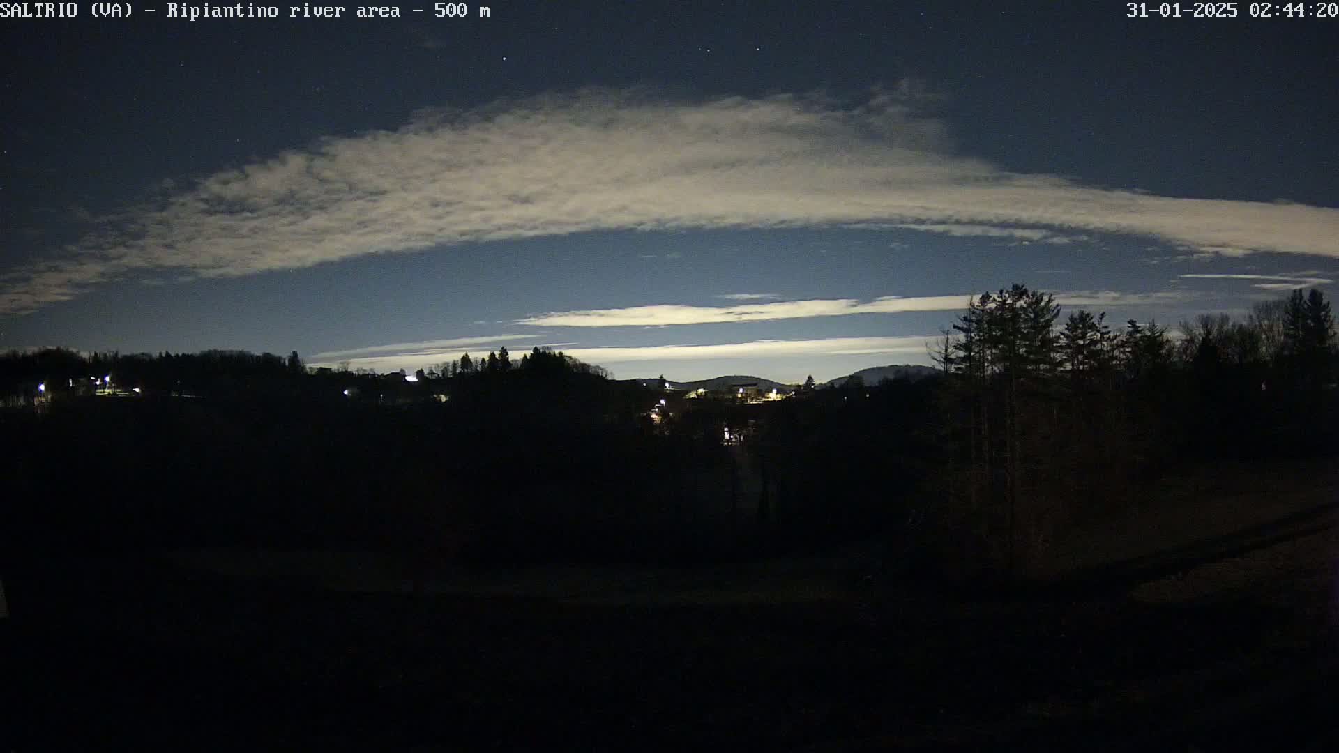 A nighttime view of a dark, silhouetted landscape under a mostly clear sky with thin, high clouds.