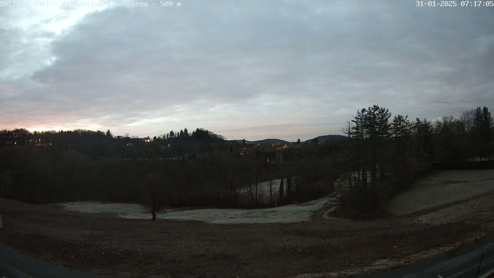 A pre-dawn view shows a mostly cloudy sky over a landscape of dark trees, a frost-covered field, and a small town in the distance.