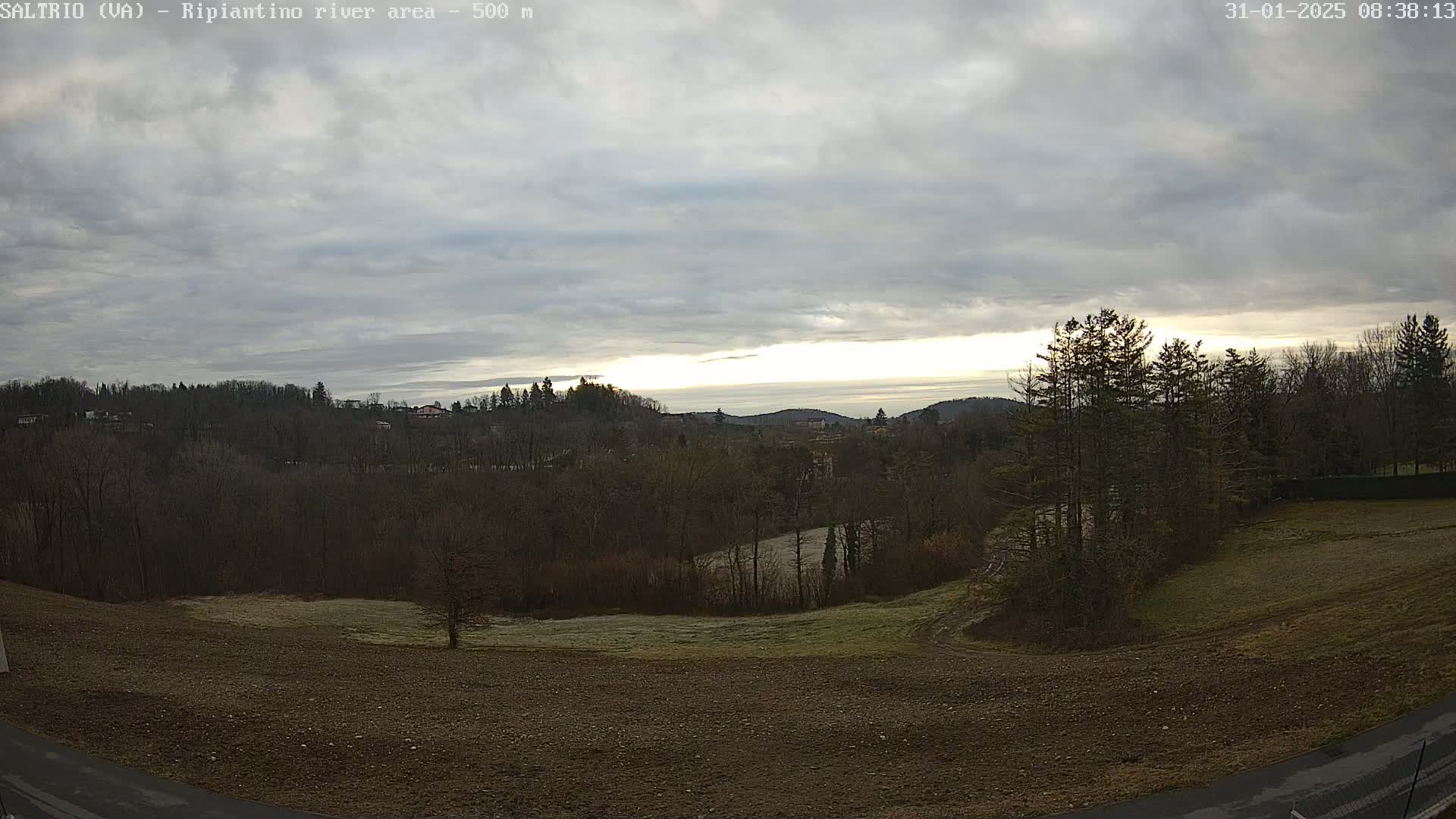 A mostly cloudy sunrise illuminates a hilly landscape of bare winter trees and a plowed field, with a small river visible in the distance.