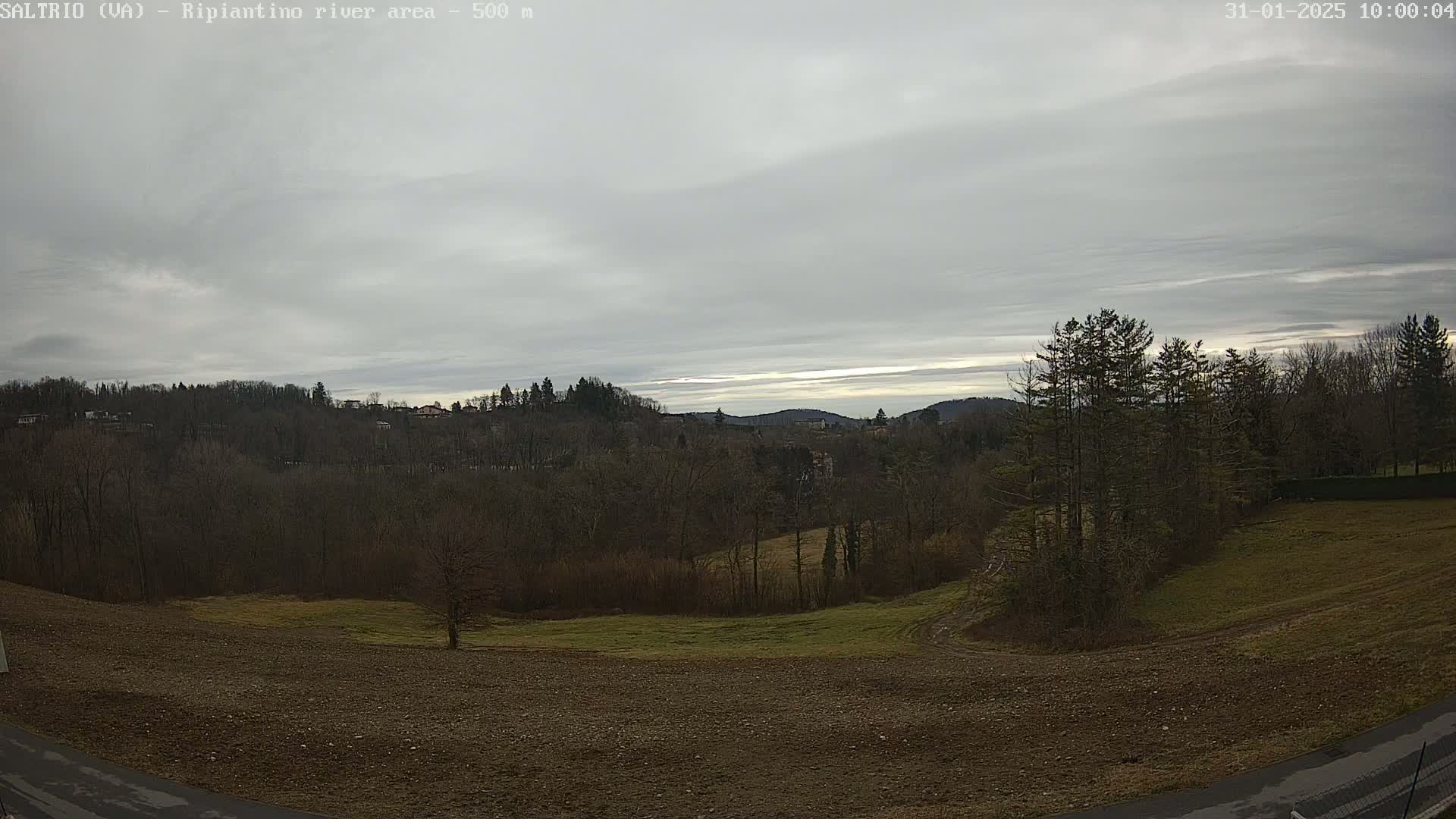 A mostly cloudy sky overlooks a plowed field and bare winter trees bordering a distant hilly landscape.