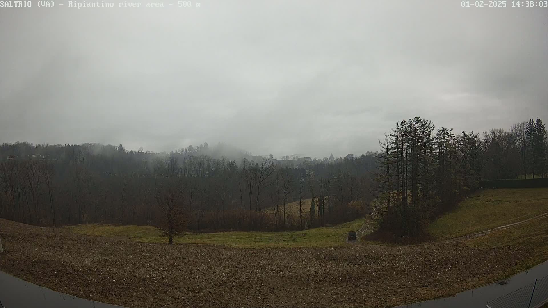 A fog-shrouded landscape reveals a hilly area covered in bare winter trees, with a small vehicle visible in a field below, under an overcast sky.