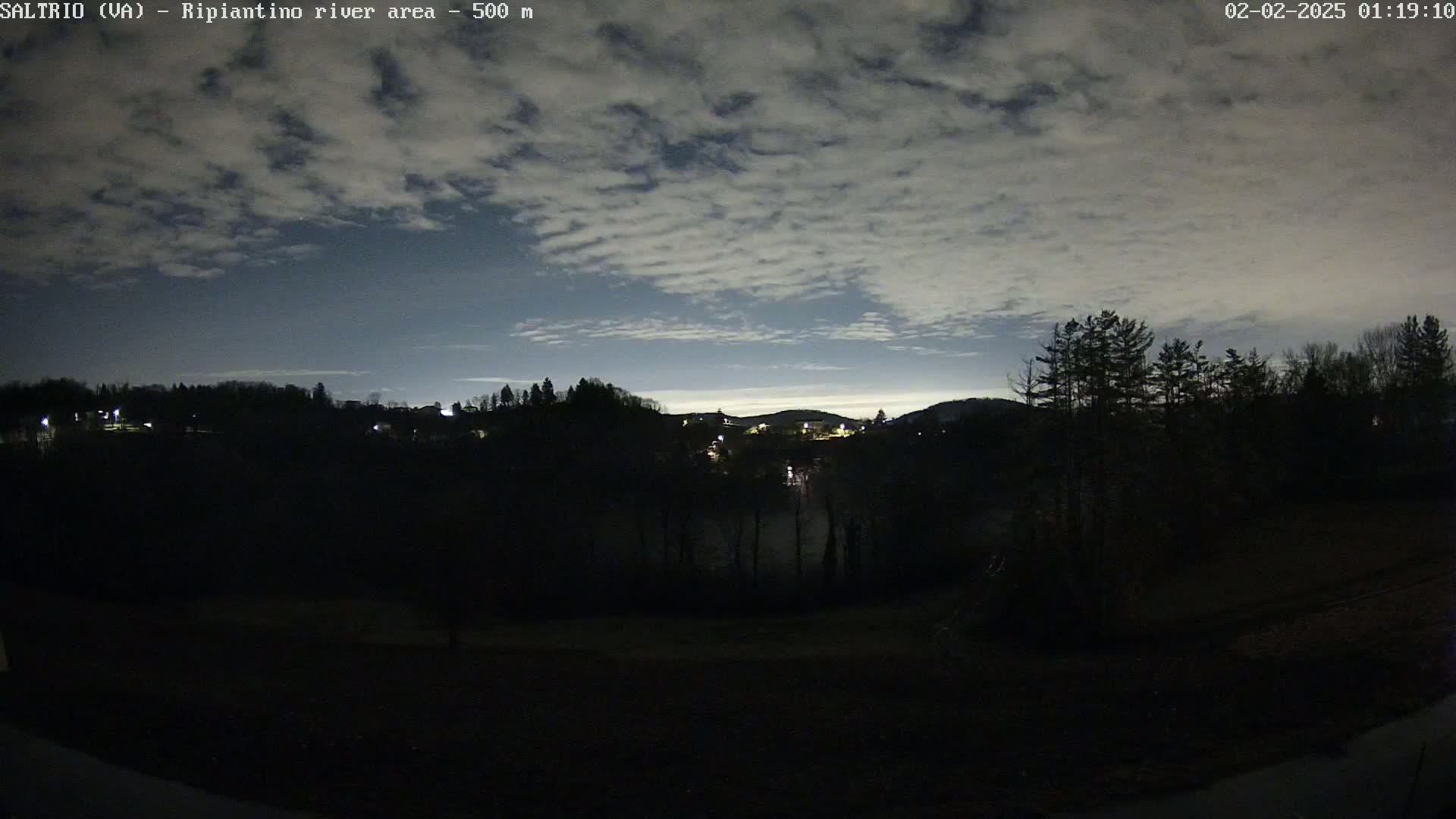 A nighttime view of a dark landscape with a river, distant lights, and a cloudy sky.