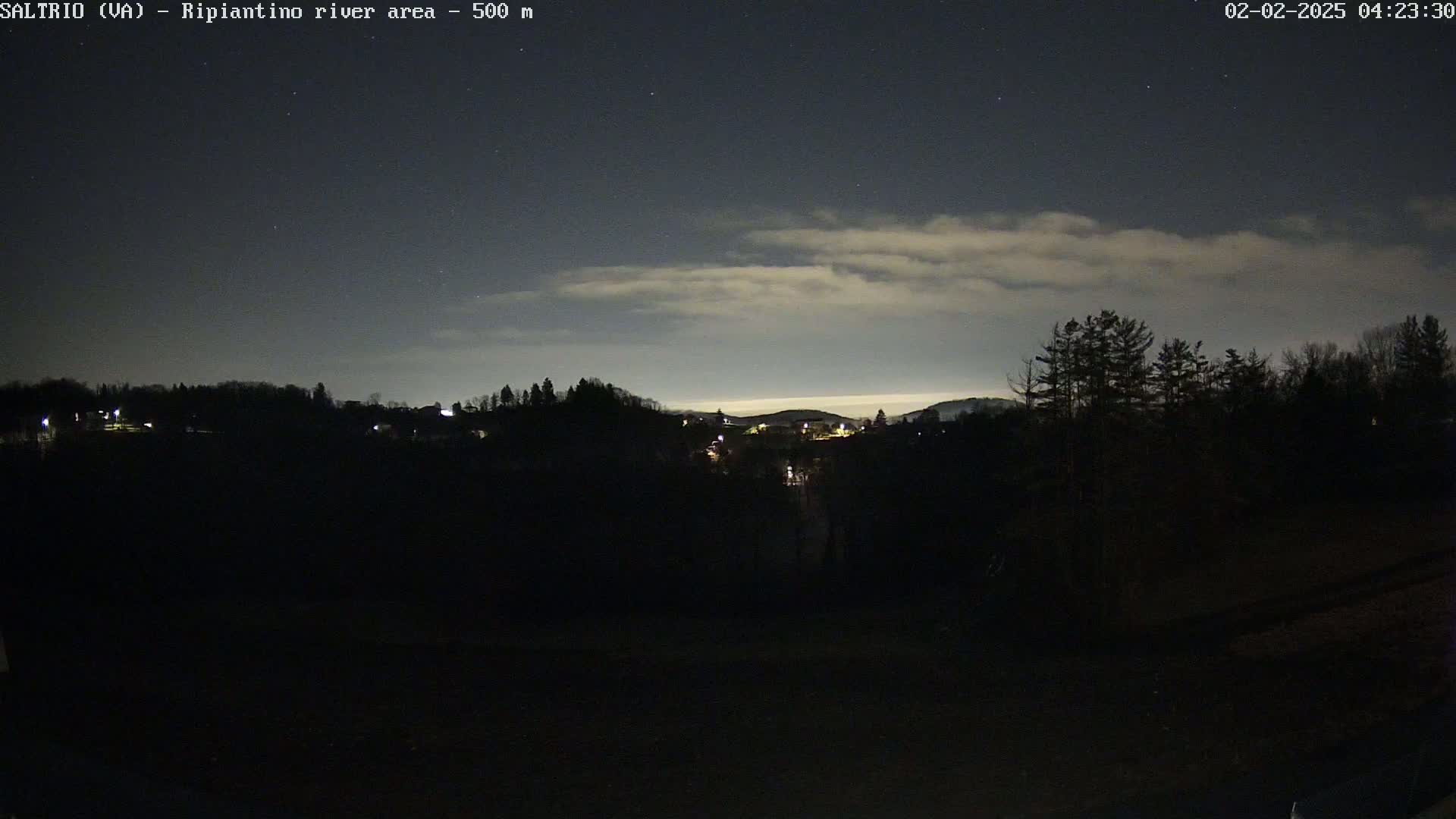 A nighttime view of a dark, hilly landscape with a few distant lights under a partly cloudy sky.