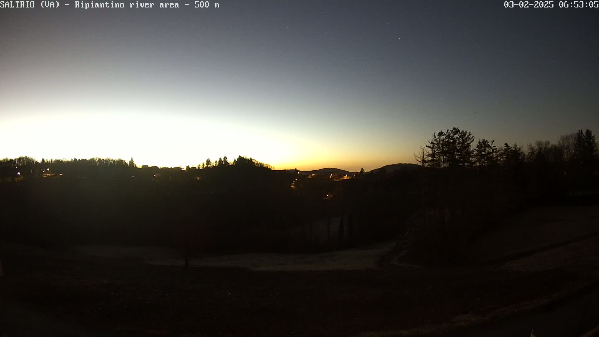 A pre-dawn sunrise illuminates a dark landscape of trees and a few distant lights, with a mostly clear sky.