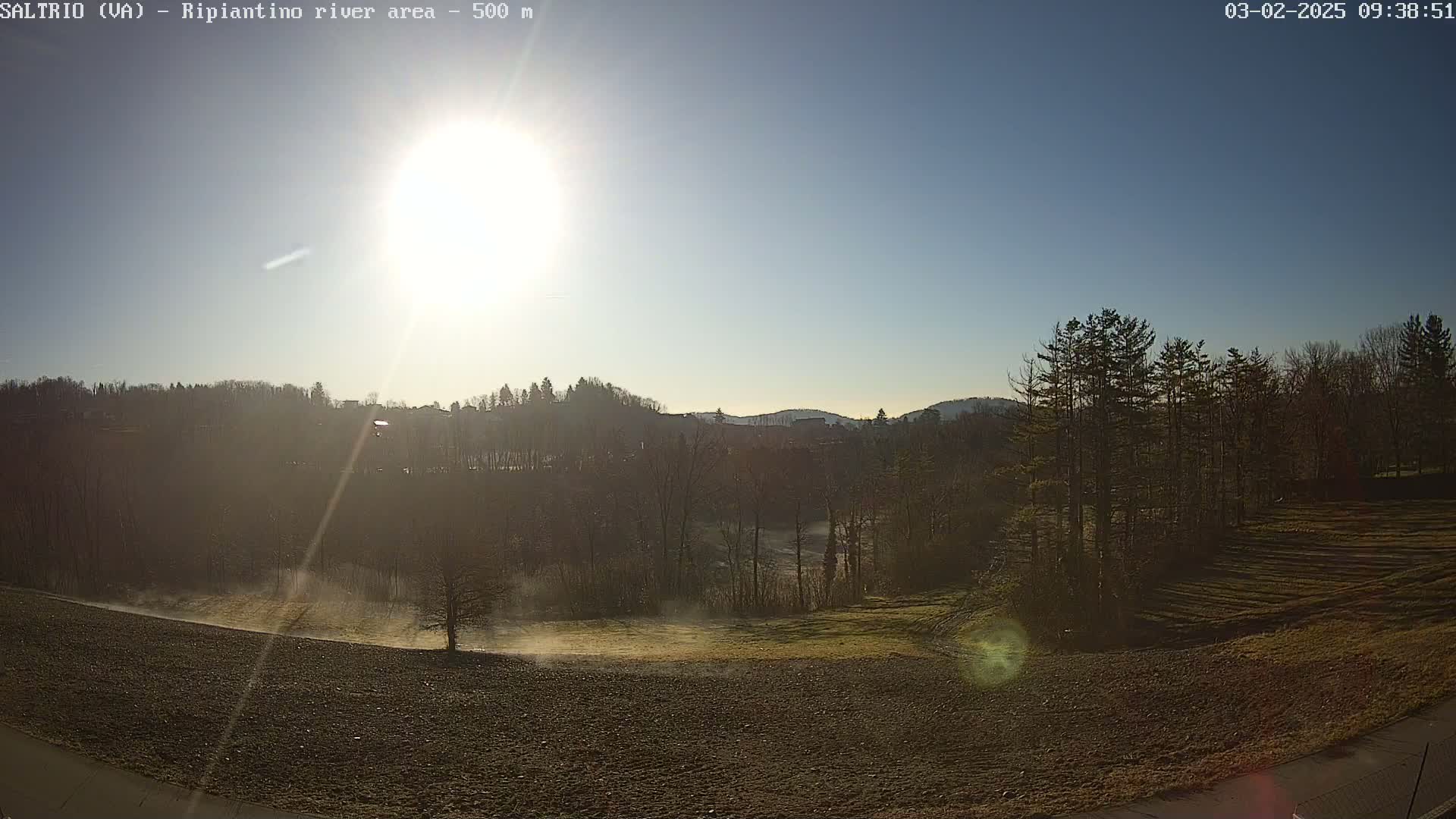 A bright sun shines in a clear blue sky over a landscape of trees, fields, and a river valley, with a light mist rising from the low-lying areas.