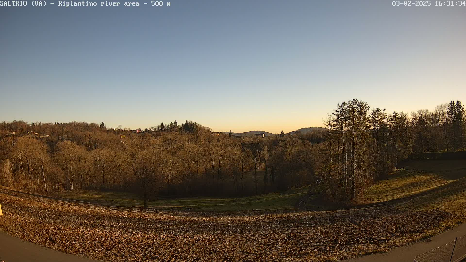 A mostly clear, twilight sky overlooks a plowed field and a hilly landscape of leafless trees.