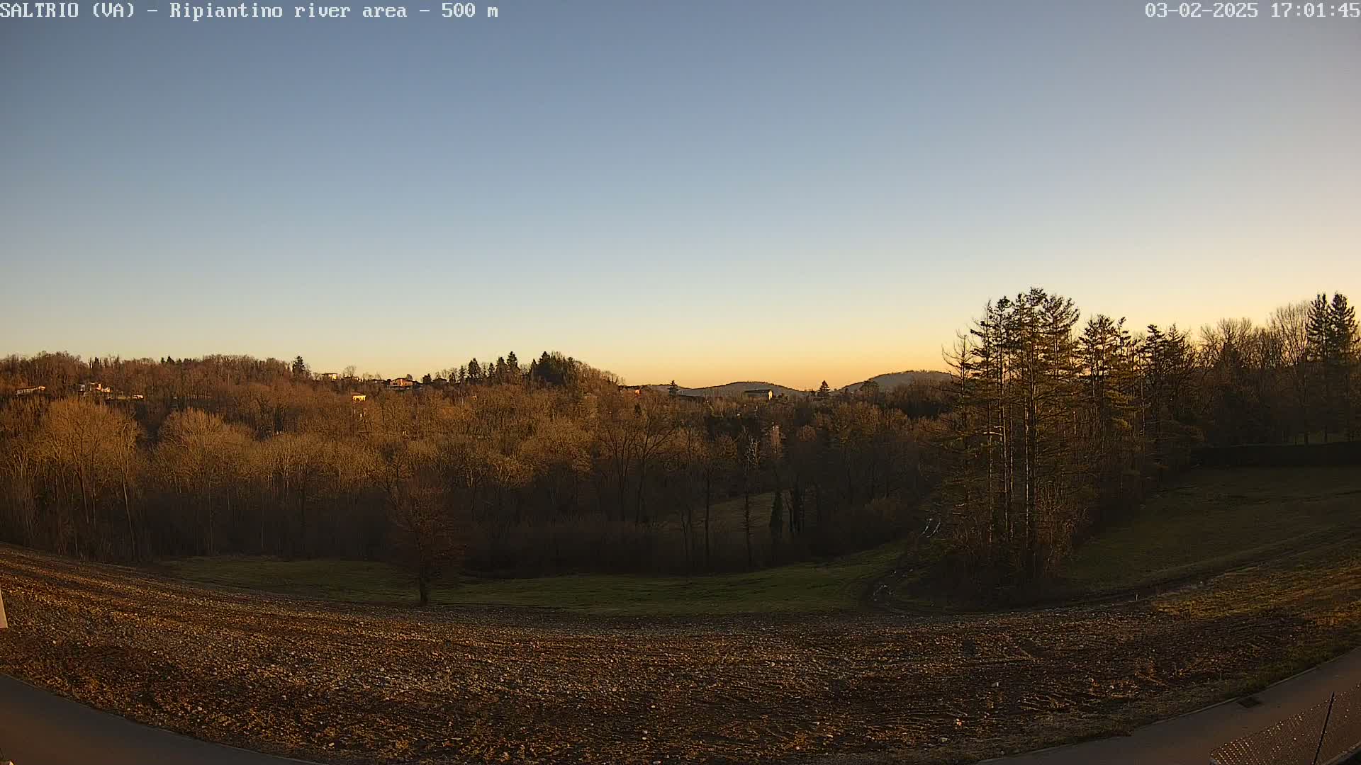 A mostly clear sunset illuminates a plowed field in the foreground, with a line of trees and hills beyond.