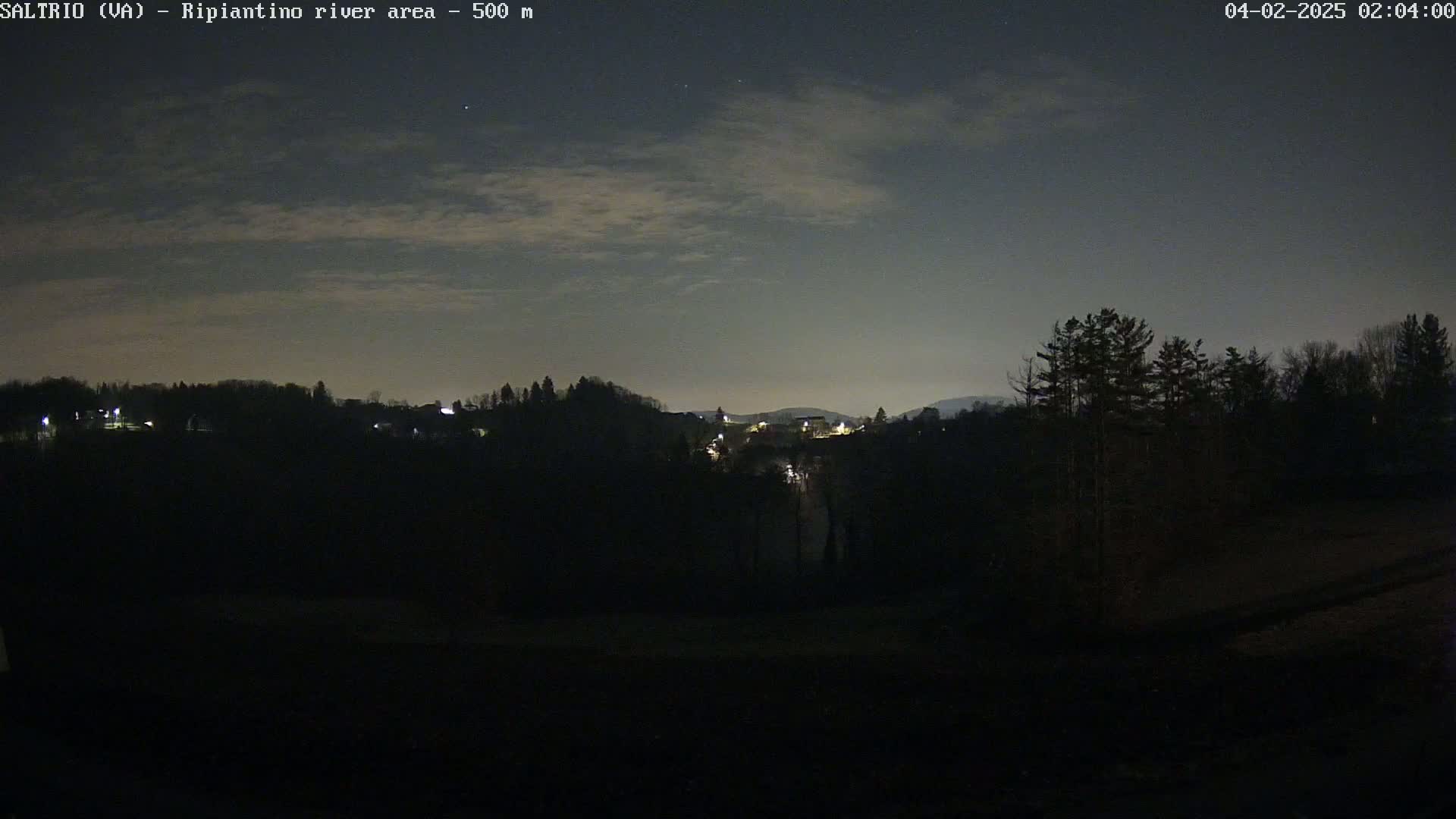 A nighttime view of a dark, hilly landscape with sparsely lit buildings and trees under a partly cloudy sky.
