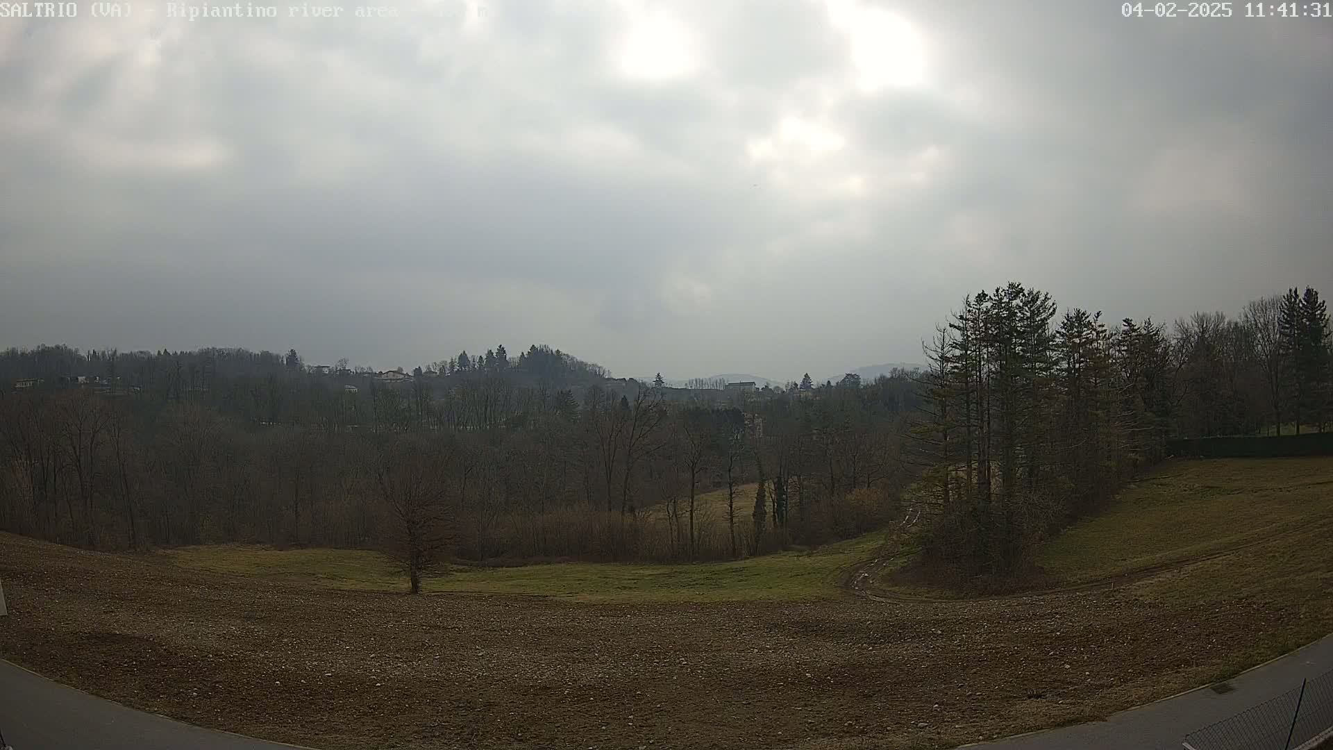 A mostly cloudy day reveals a view of bare, winter trees and brown fields, with a distant line of hills and buildings visible in the background.
