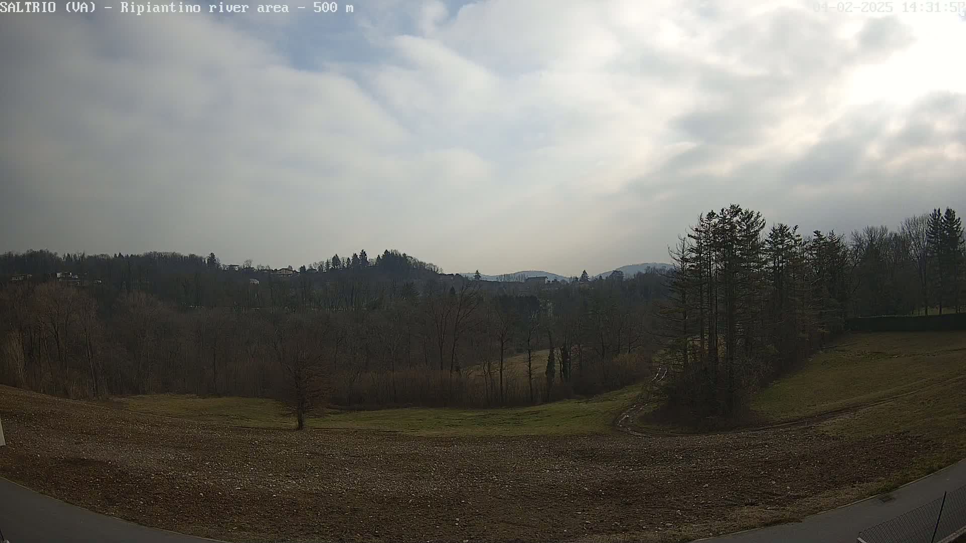 A mostly cloudy sky overlooks a landscape of bare trees, plowed fields, and distant hills.