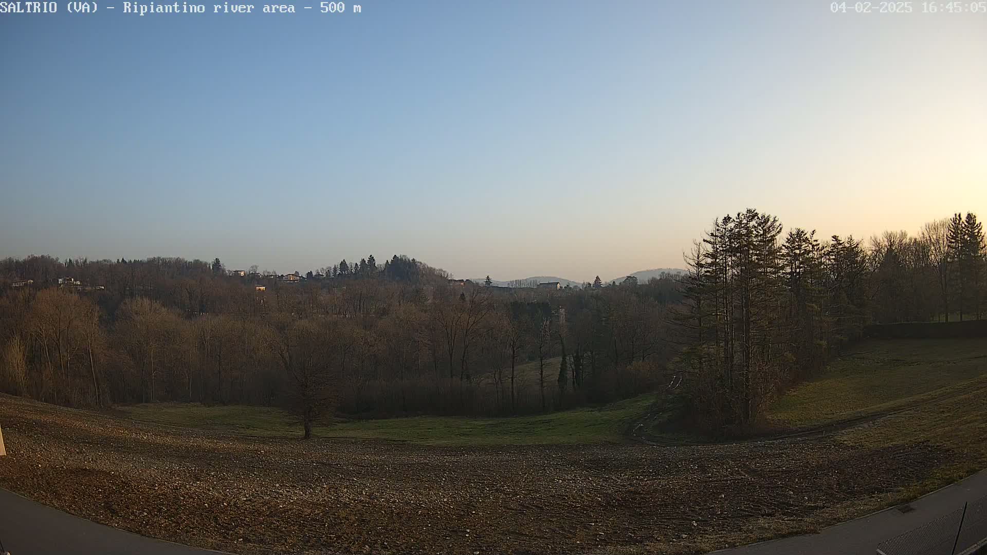 A mostly clear, pale-blue sky illuminates a hilly landscape of bare trees and tilled fields at sunset.