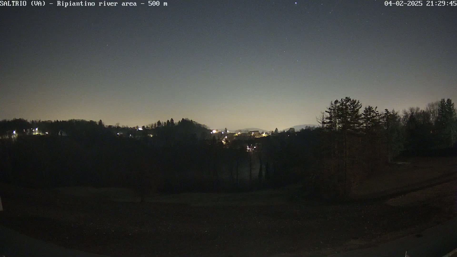 A nighttime view of a dark landscape with a few lights visible in the distance under a mostly clear, starry sky.