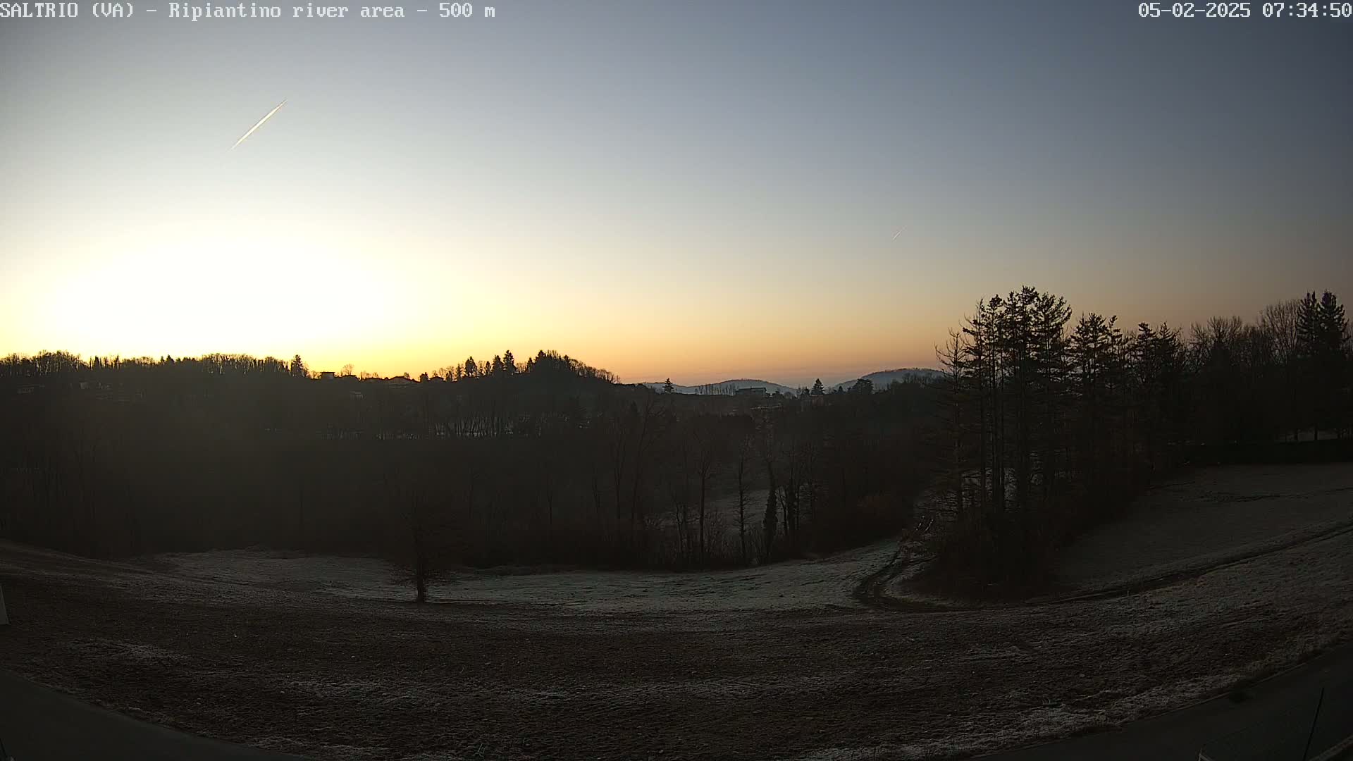 A sunrise illuminates a frost-covered field and silhouetted trees, with a contrail visible in the pale sky.