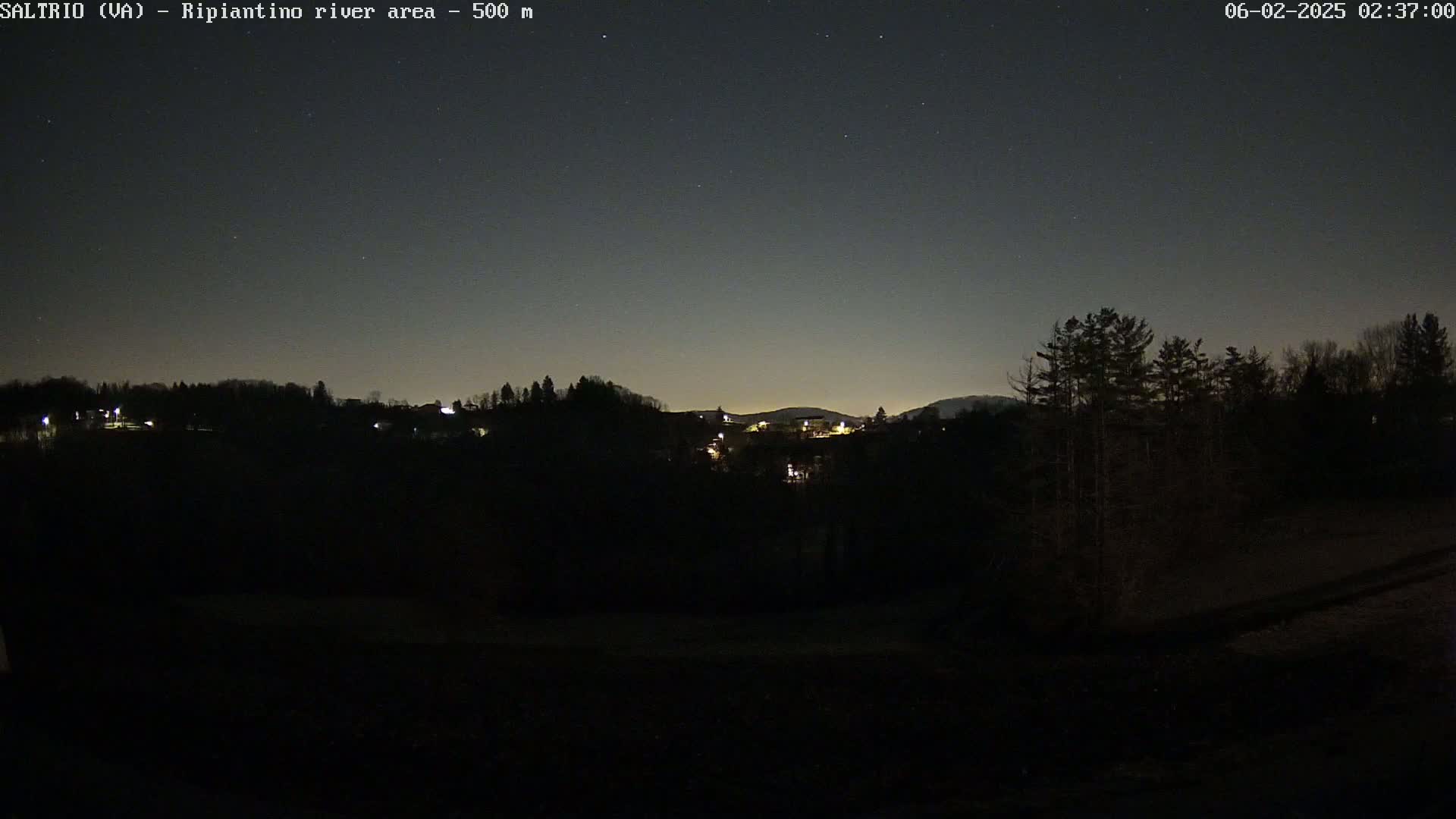 A nighttime view of a dark, hilly landscape with sparsely lit buildings and trees under a clear, starry sky.