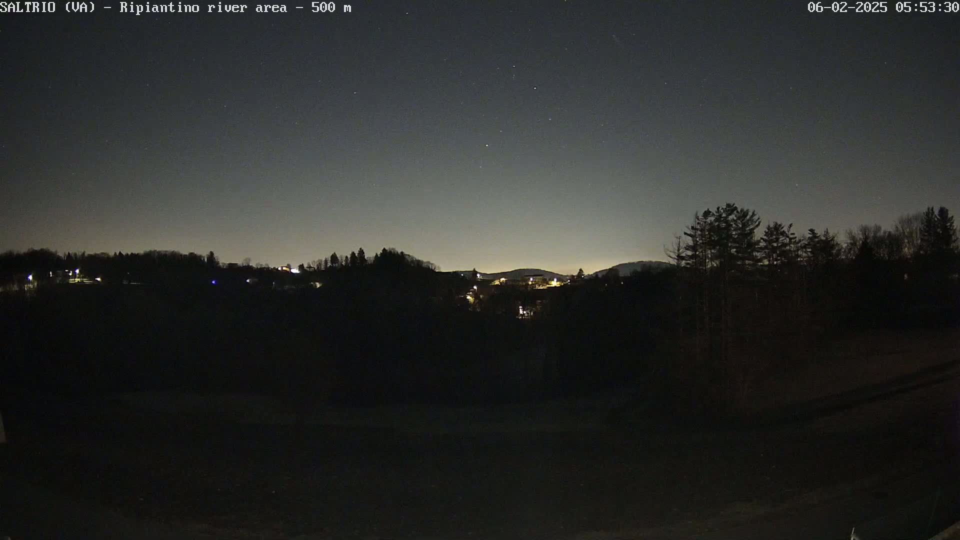 A nighttime view of a dark, hilly landscape with a few lights visible in the distance under a clear, starry sky.