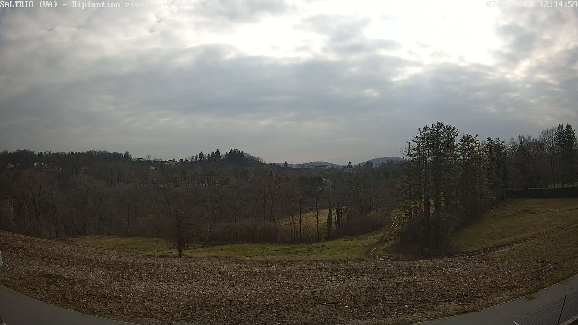 A mostly cloudy day reveals a view of a plowed field, bare winter trees, and distant hills.