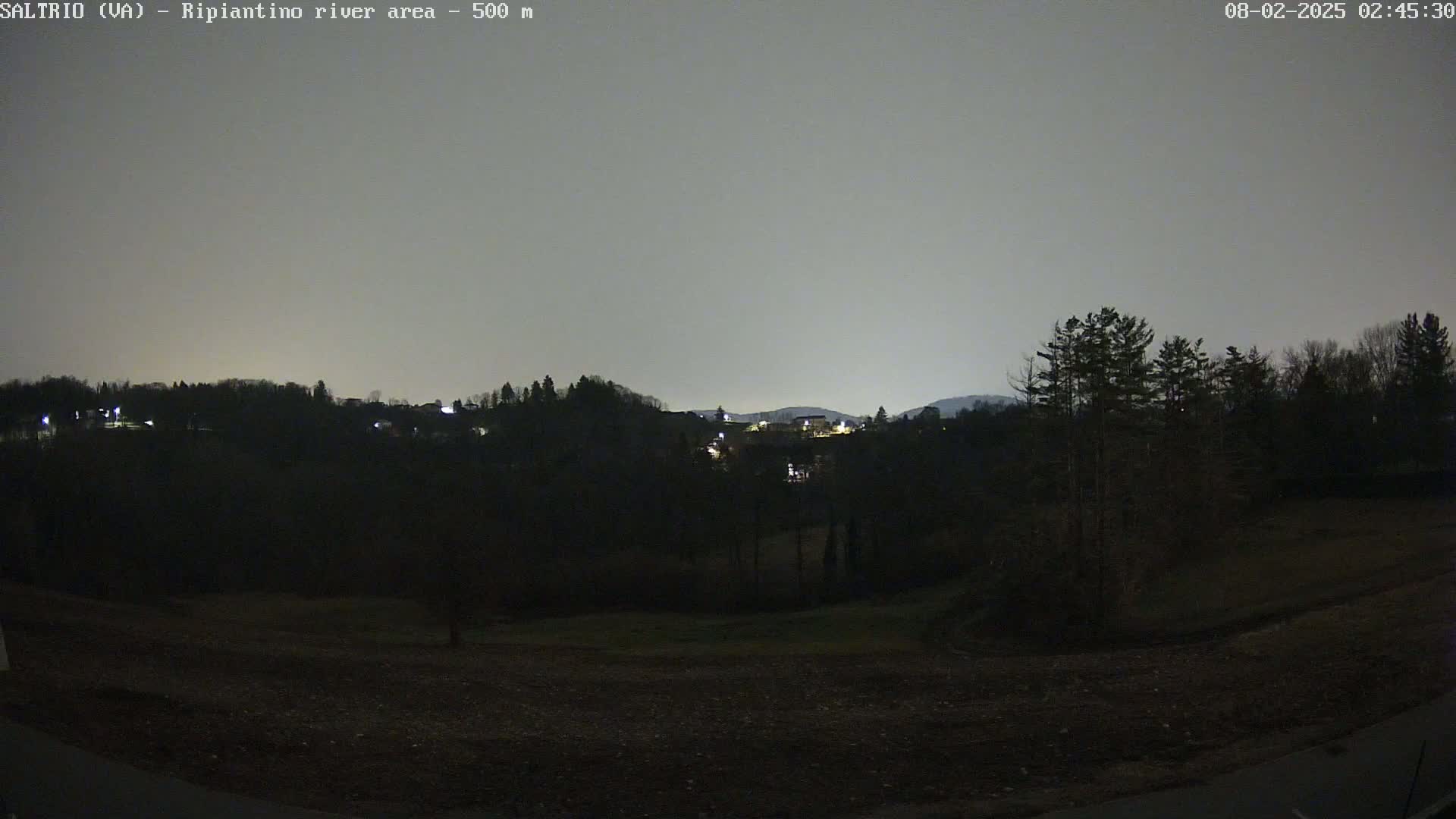 A nighttime view of a dark, hilly landscape with sparsely lit buildings in the distance under a gray sky.