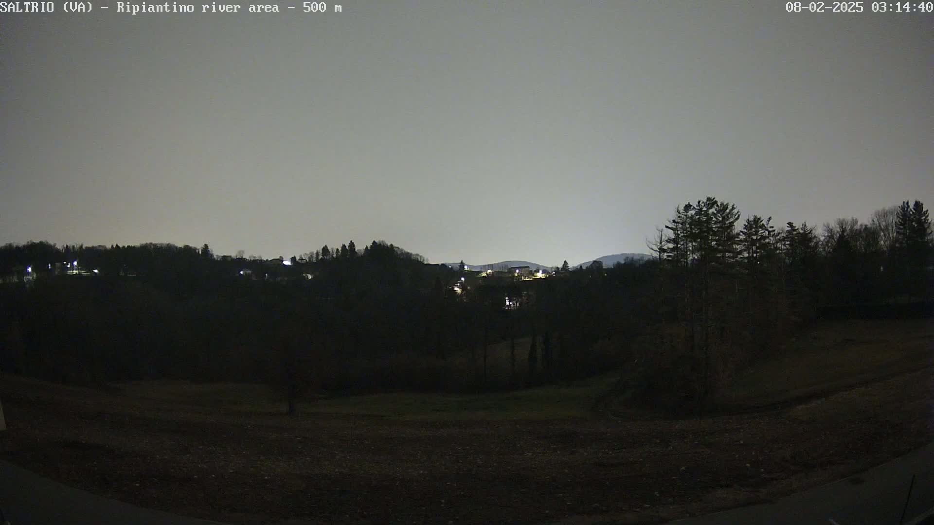 A nighttime view of a dark, mostly treeless landscape with a few lights visible in the distance under a light gray sky.
