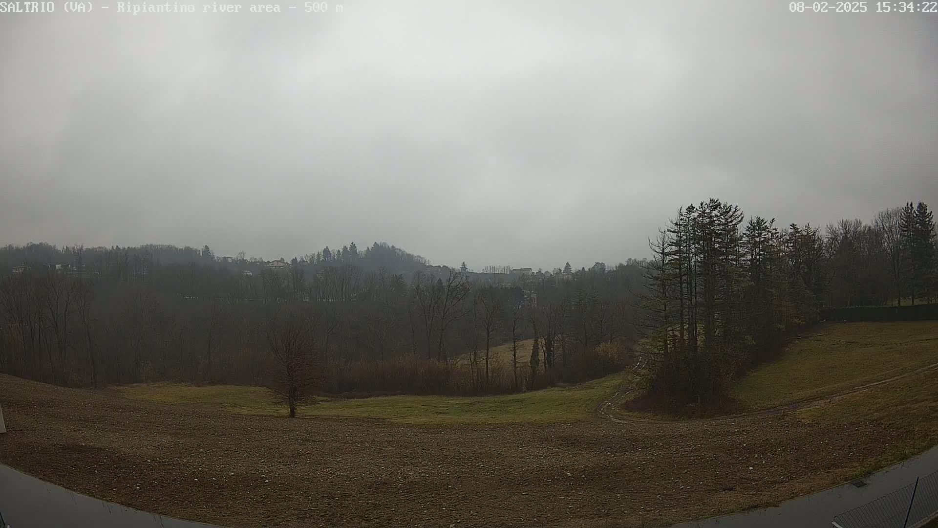 A gray, overcast day reveals a view of a plowed field, sparsely vegetated areas, and a line of trees bordering a wooded hillside in the distance.