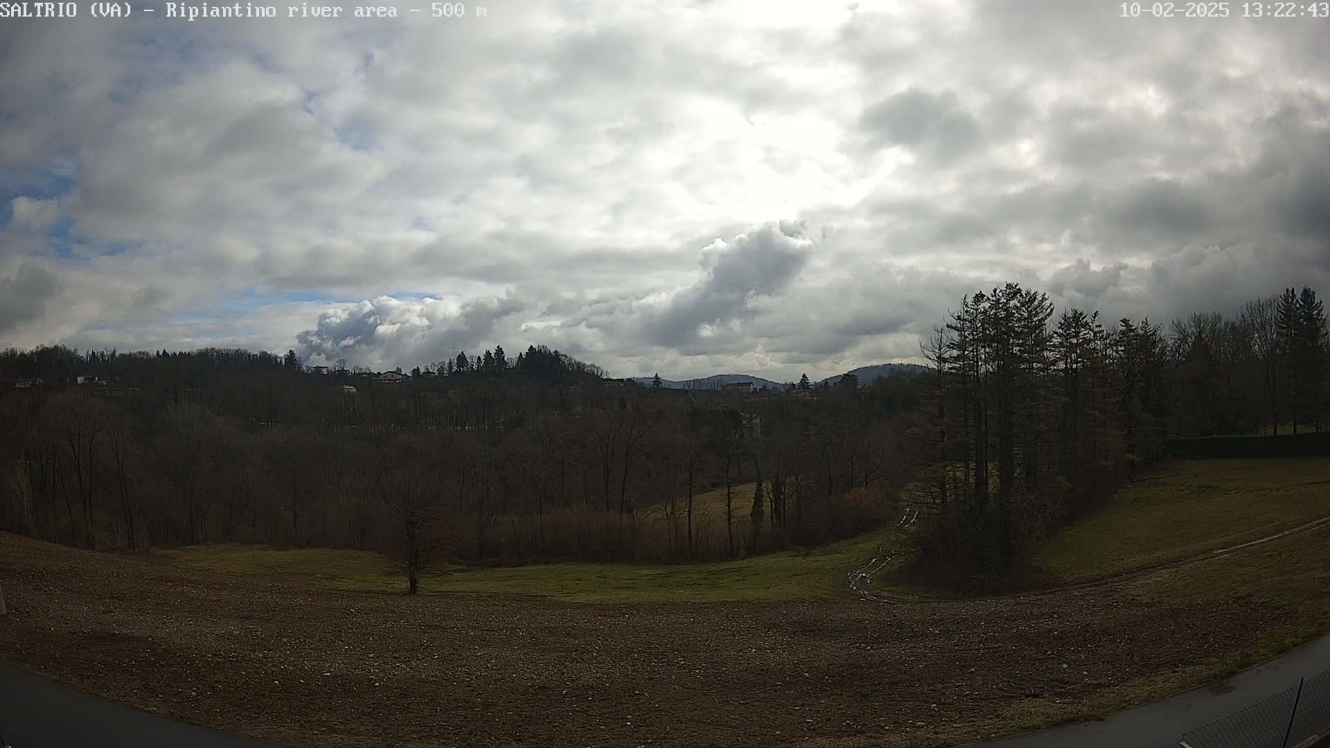 A partly cloudy sky overlooks a mostly barren landscape of dark trees, patches of green grass, and brown, tilled earth.