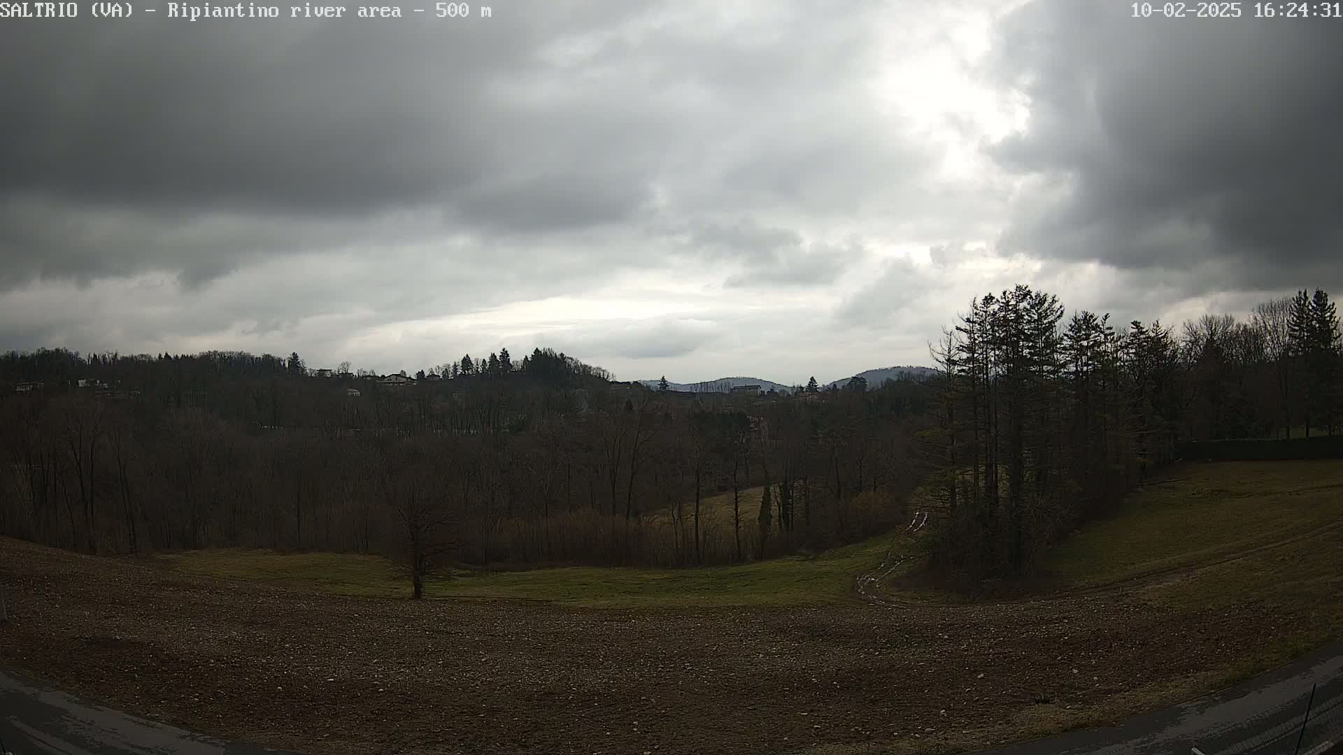 A mostly cloudy sky hangs over a mostly bare, hilly landscape with patches of brown and green fields and dark-colored trees.