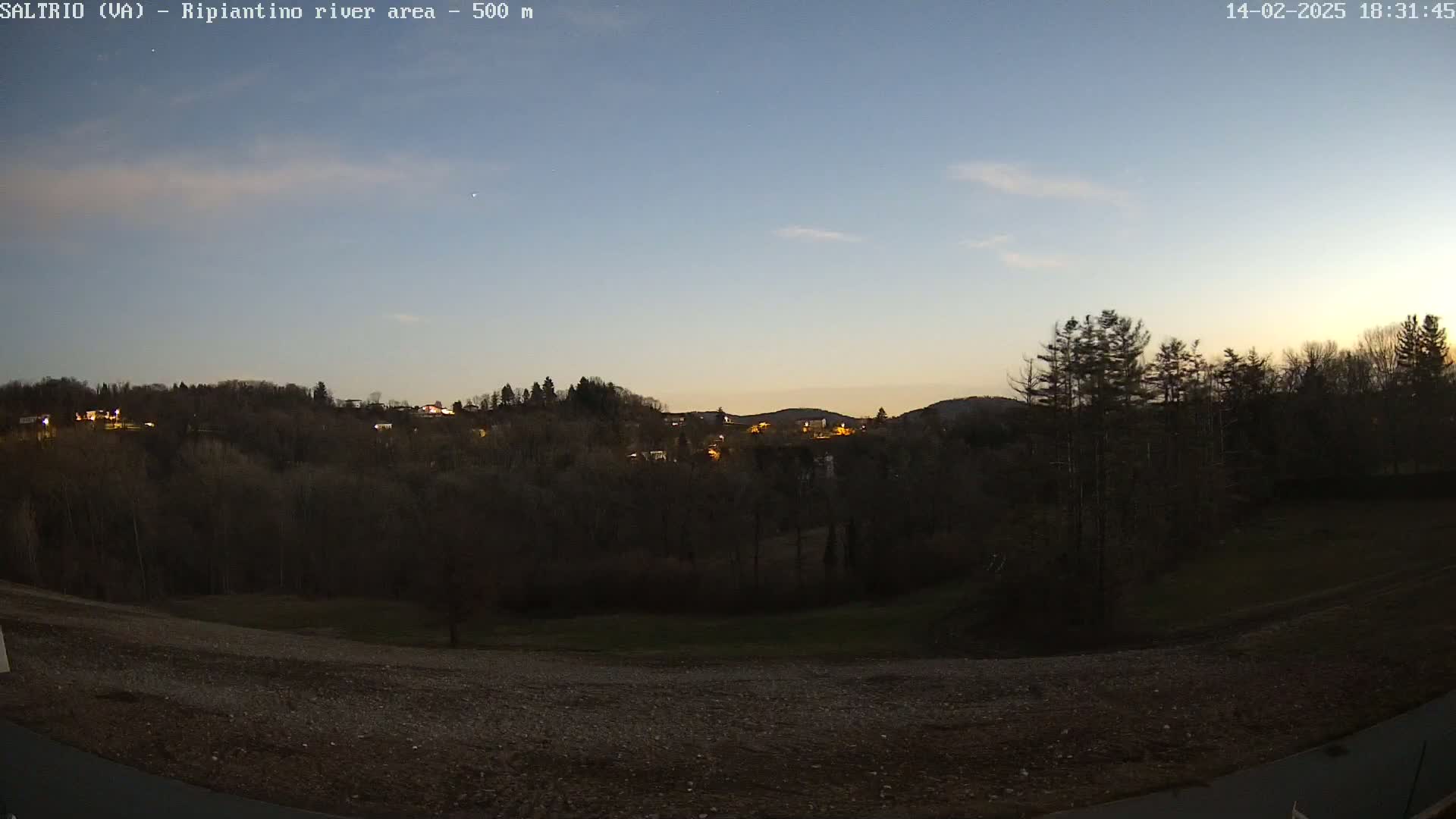 A twilight view of a hilly landscape with sparse, dark trees, a few distant lights, and a mostly clear sky.