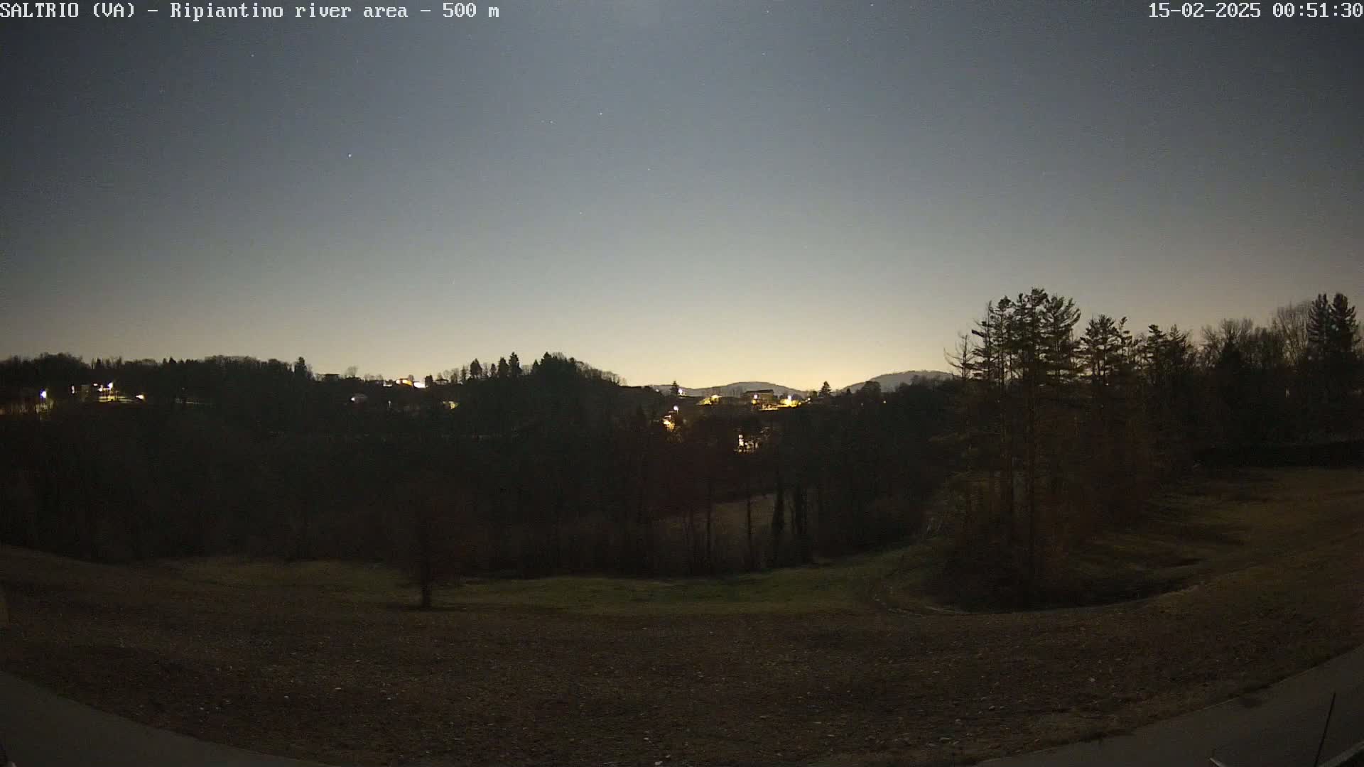 A nighttime view of a dark, tree-lined hillside with a few scattered lights, under a twilight sky.