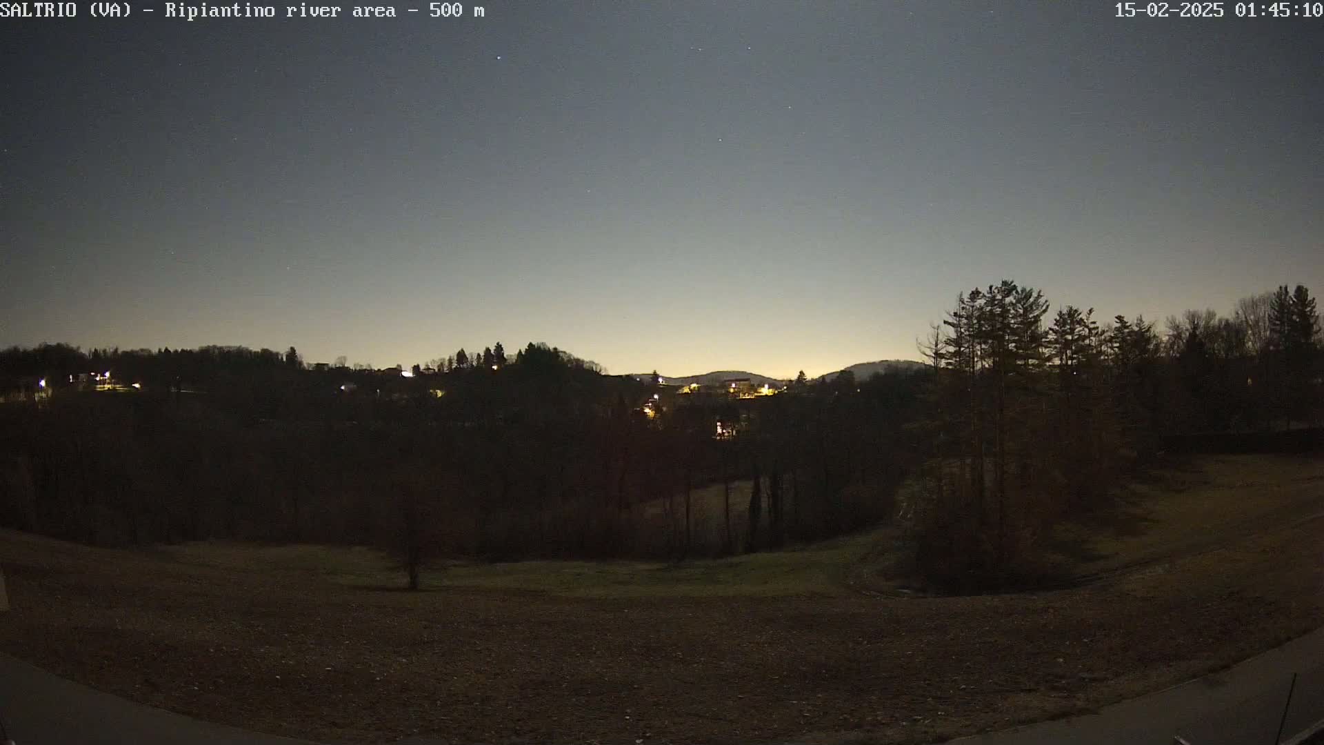 A nighttime view of a dark, hilly landscape with sparsely lit buildings in the distance under a clear, twilight sky.