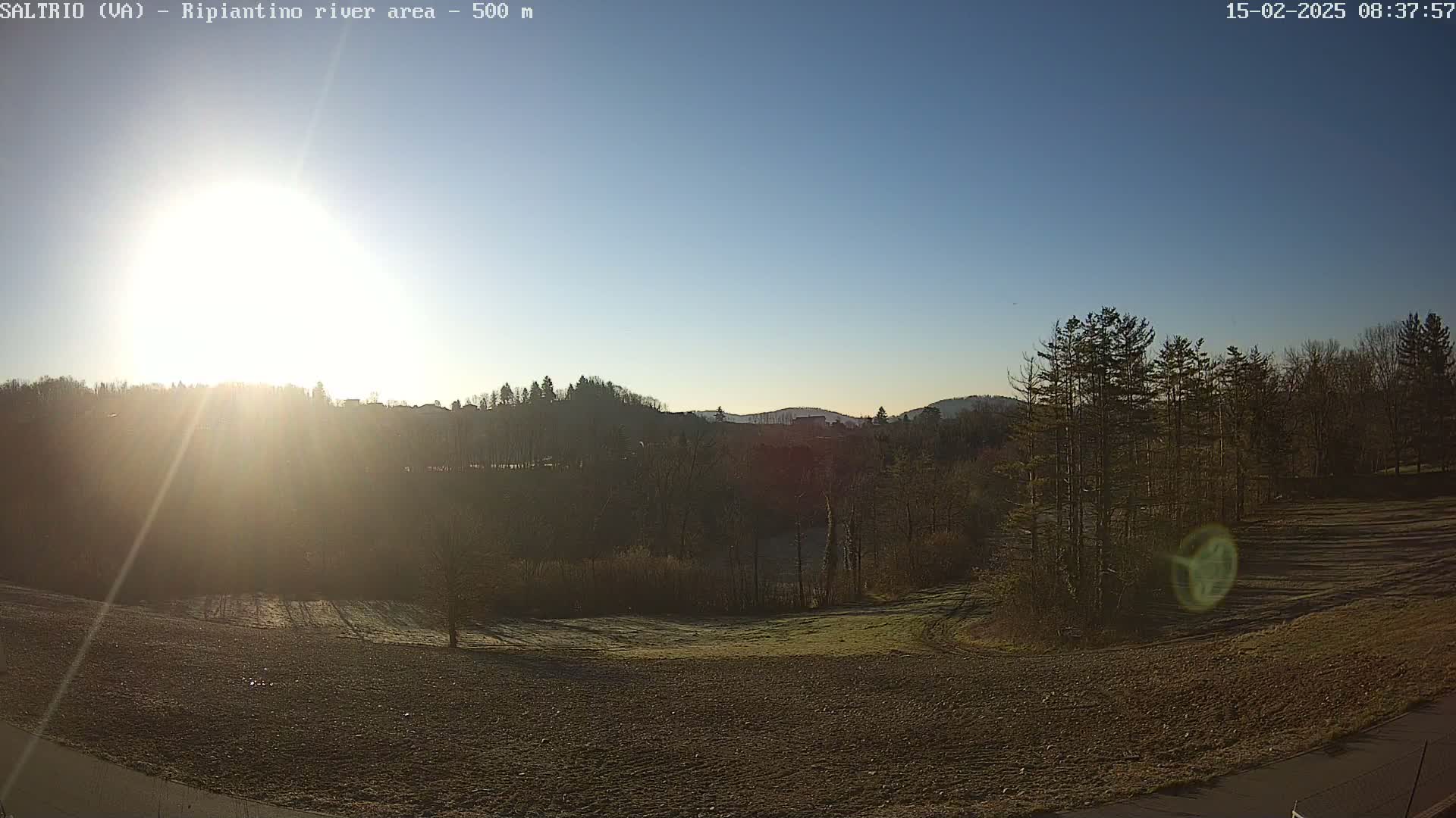 A bright sun shines over a plowed field and trees on a clear, cold morning.