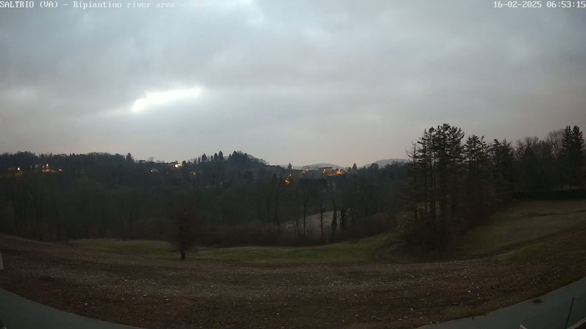 A dark, overcast sky hangs over a hilly landscape of dark green trees and a plowed field, with a few lights visible in the distance.