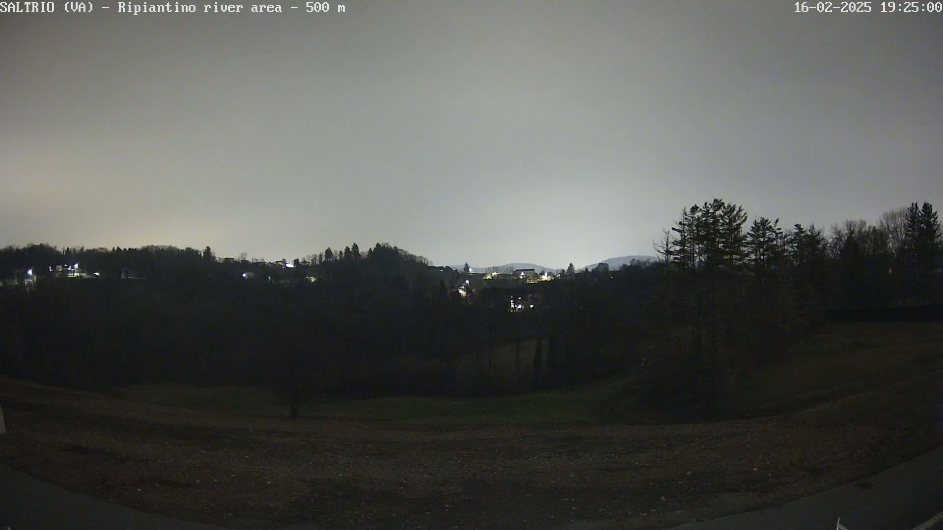 A nighttime view of a dark, hilly landscape with sparsely lit buildings in the distance under a gray, overcast sky.