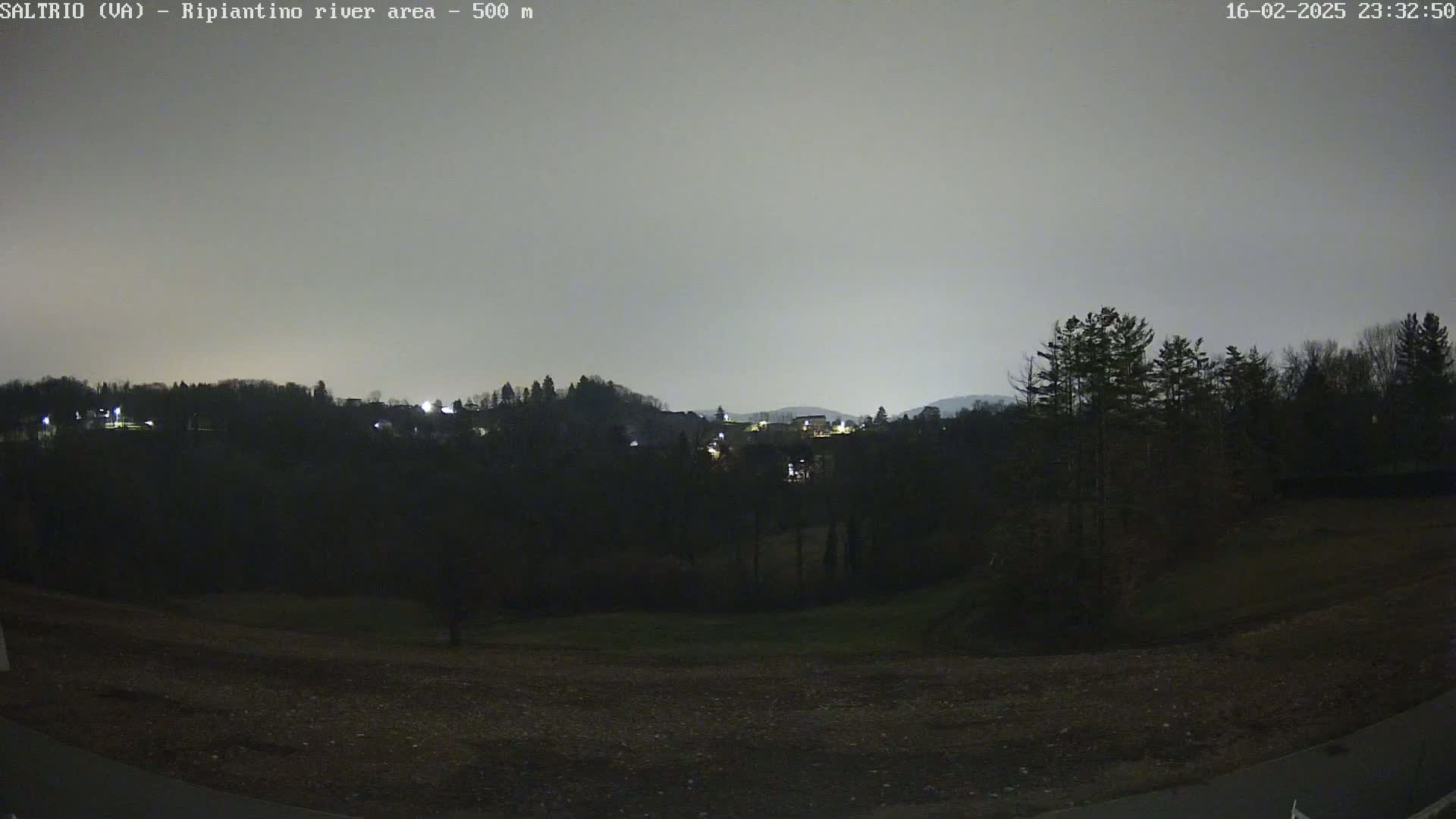 A nighttime view of a dark, hilly landscape with sparse, dimly lit buildings in the distance under a cloudy, gray sky.