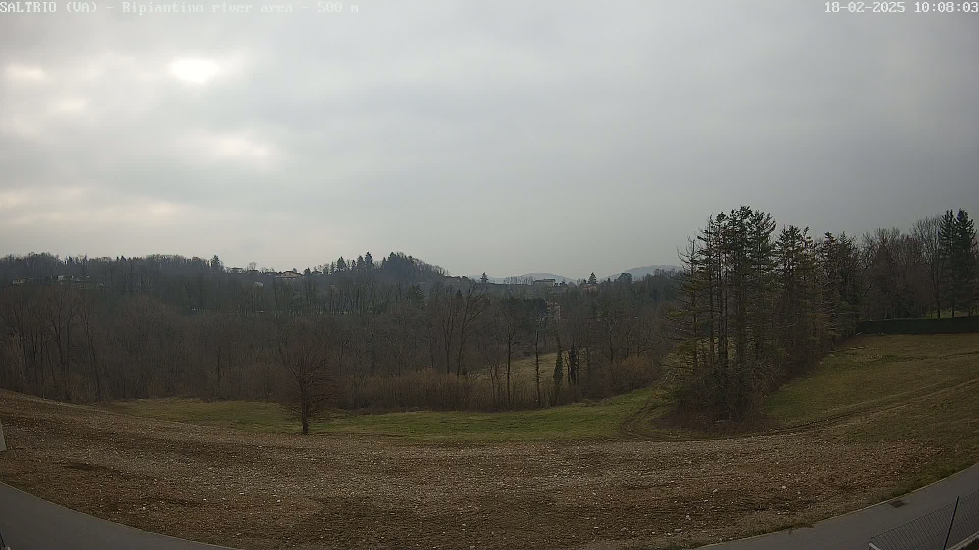 A mostly cloudy day reveals a view of a plowed field, bare winter trees, and distant hills.