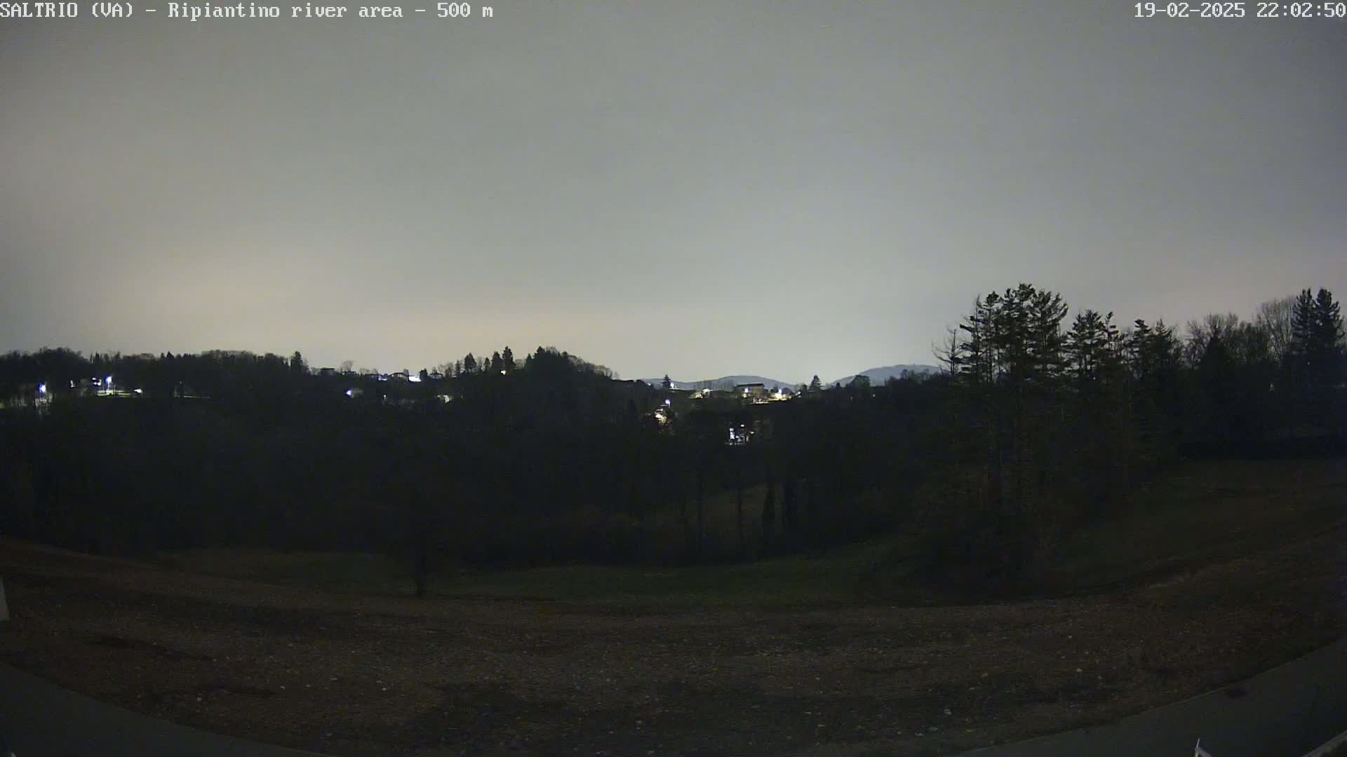 A nighttime view of a dark, hilly landscape with sparsely lit buildings in the distance under a cloudy, gray sky.