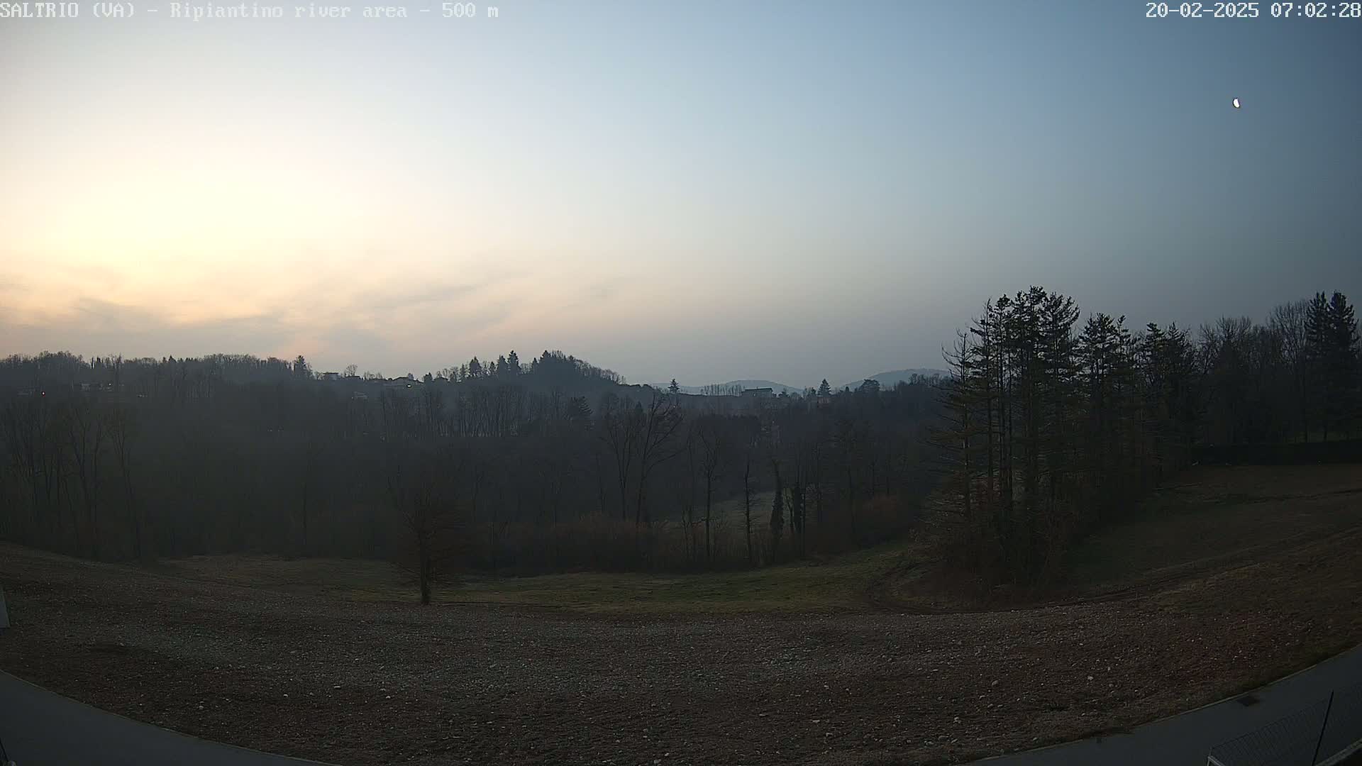A hazy sunrise illuminates a hilly landscape of dark trees and bare fields, under a pale blue sky.