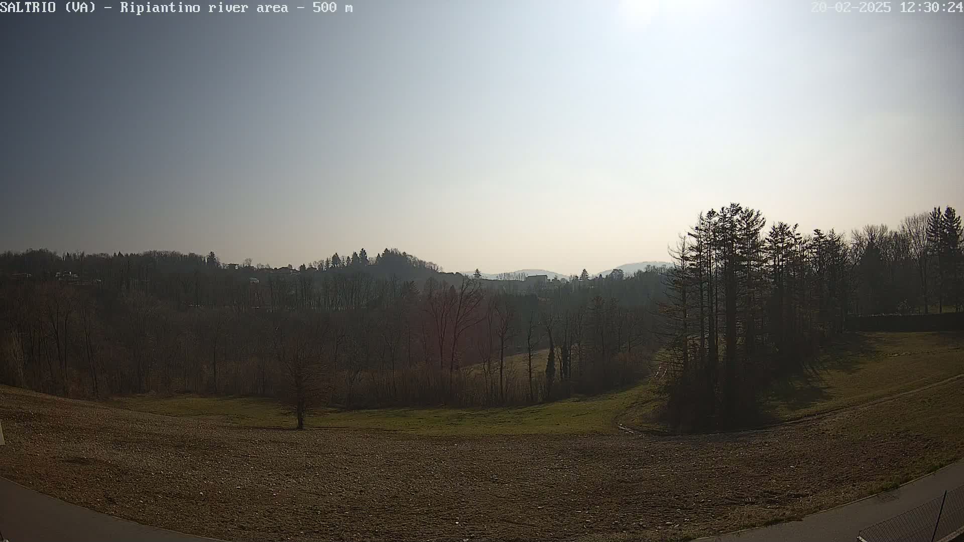 A sunny day reveals a treeline and hills in the distance, with a grassy field and bare, brown earth in the foreground.