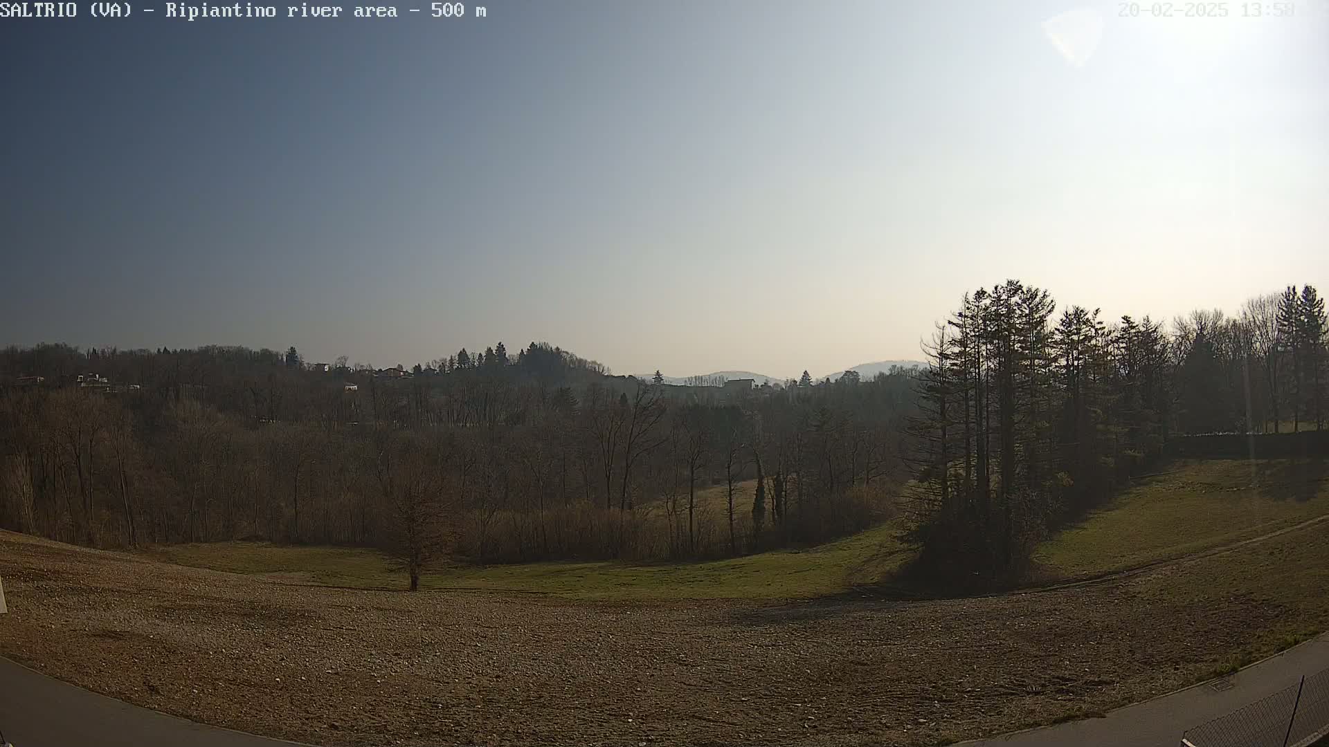 A mostly sunny, hazy day reveals a view of bare winter trees and a sparsely vegetated field sloping gently down towards a distant line of hills.