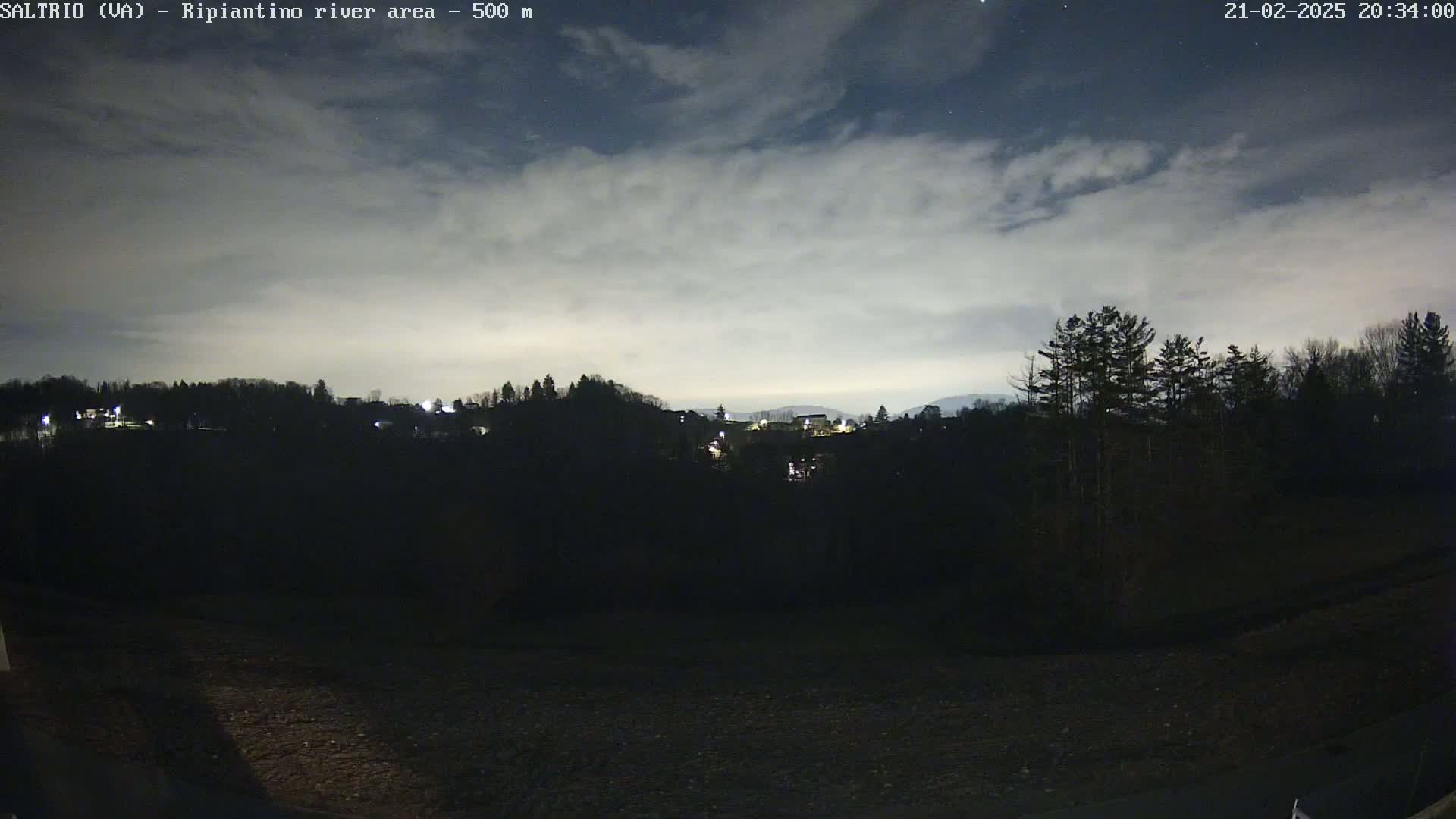 A nighttime view of a dark, tree-lined hillside with a few distant lights, under a partly cloudy sky.