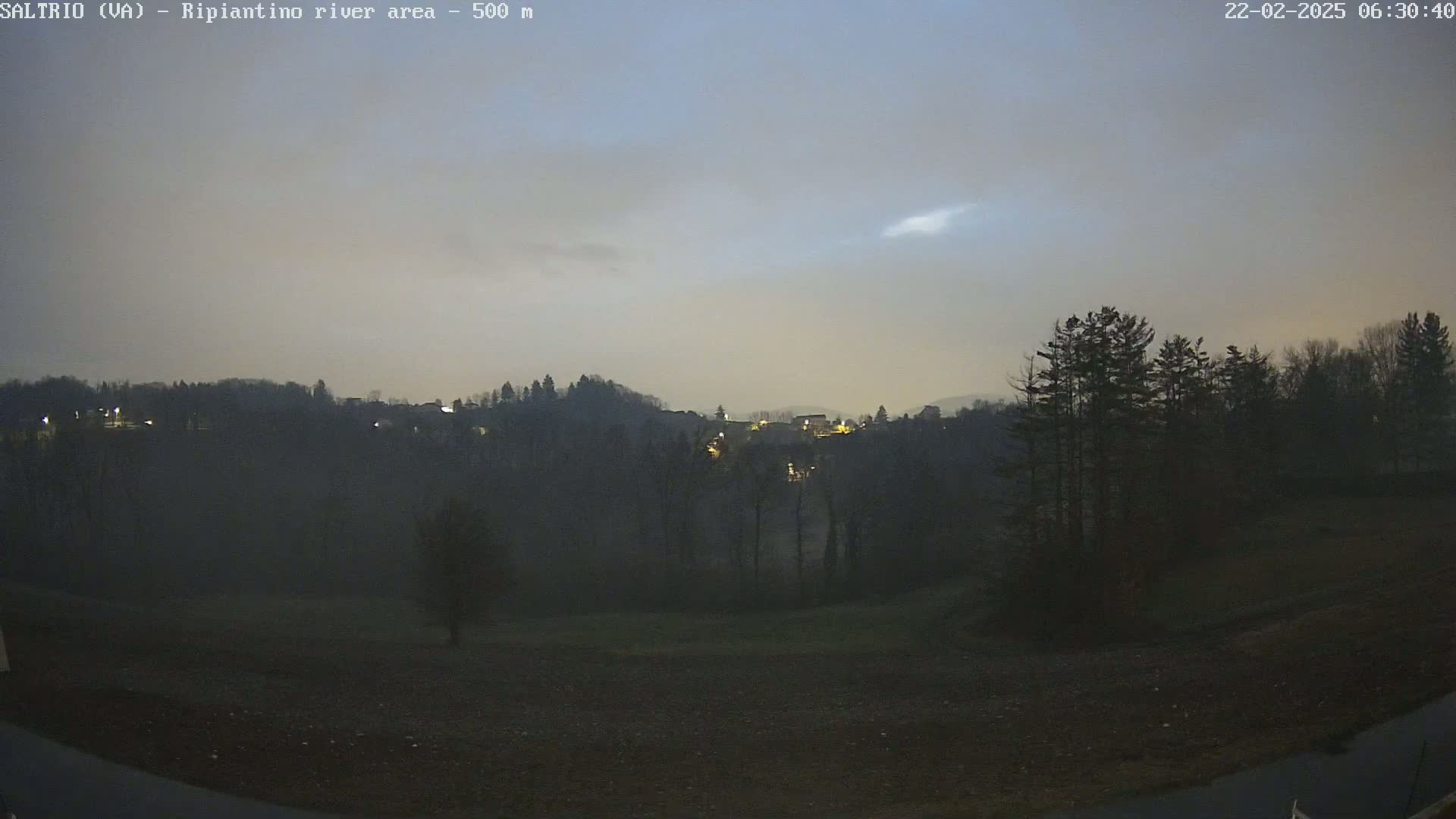 A dark, pre-dawn view of a hilly landscape with a few lights visible in the distance, under a hazy, light gray sky.