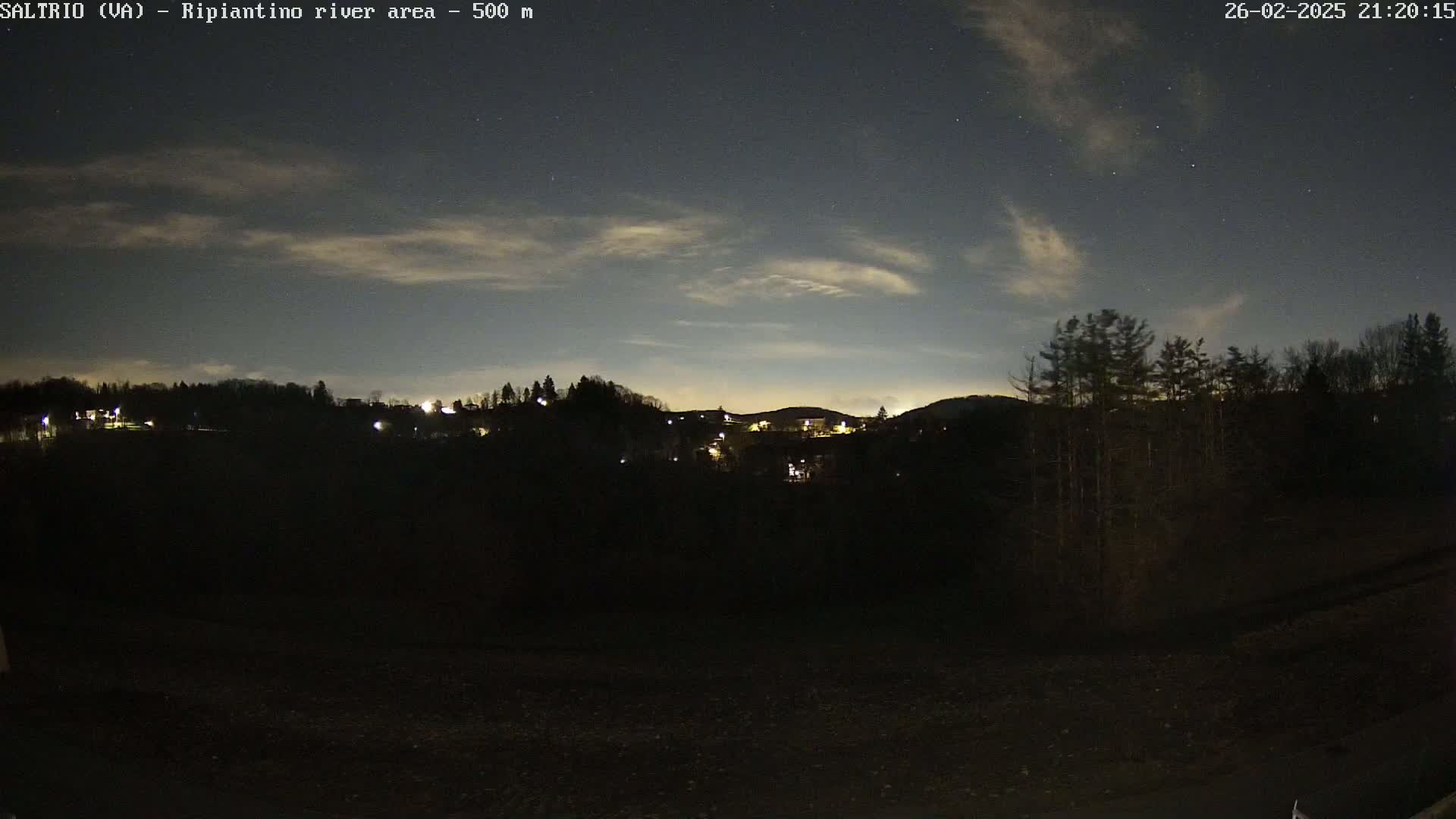 A nighttime view of a sparsely lit town nestled in a dark, tree-covered hill under a slightly cloudy night sky.