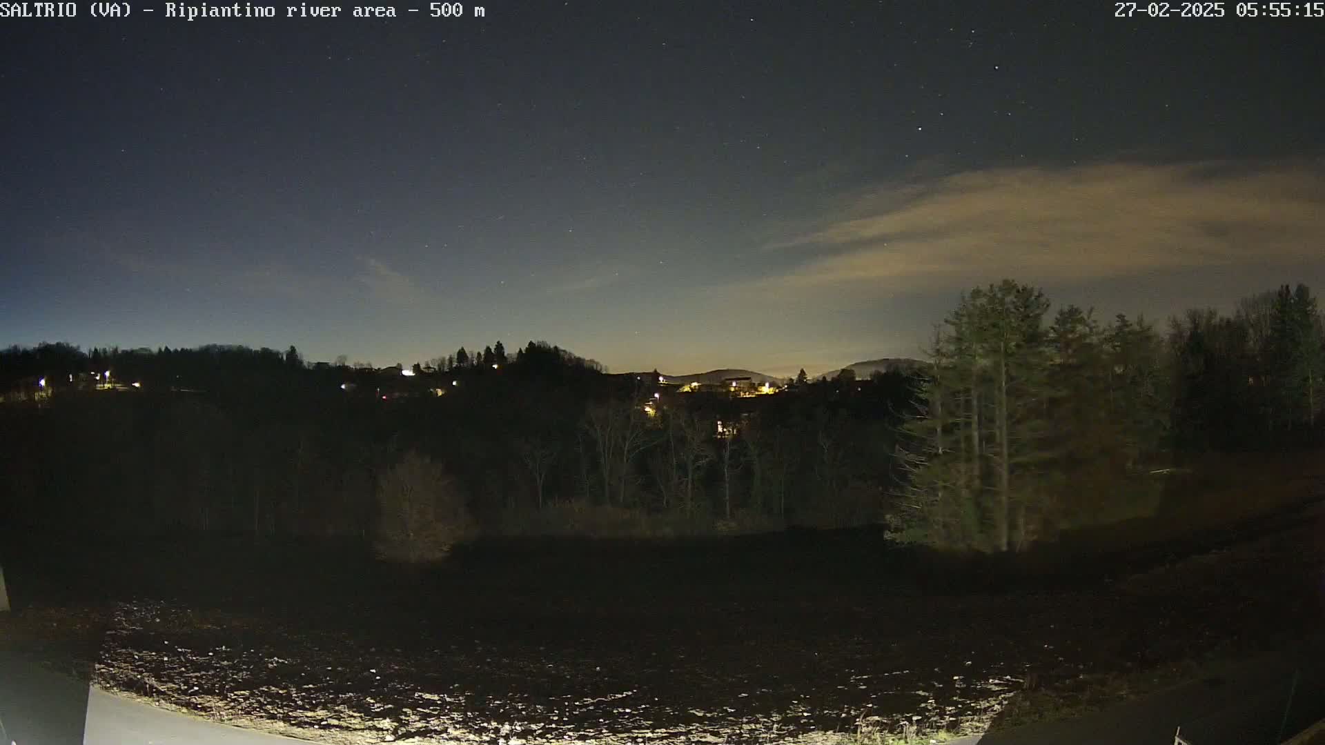 A nighttime view of a dark, tree-lined hillside with a few scattered lights, under a mostly clear sky with some faint clouds and visible stars.