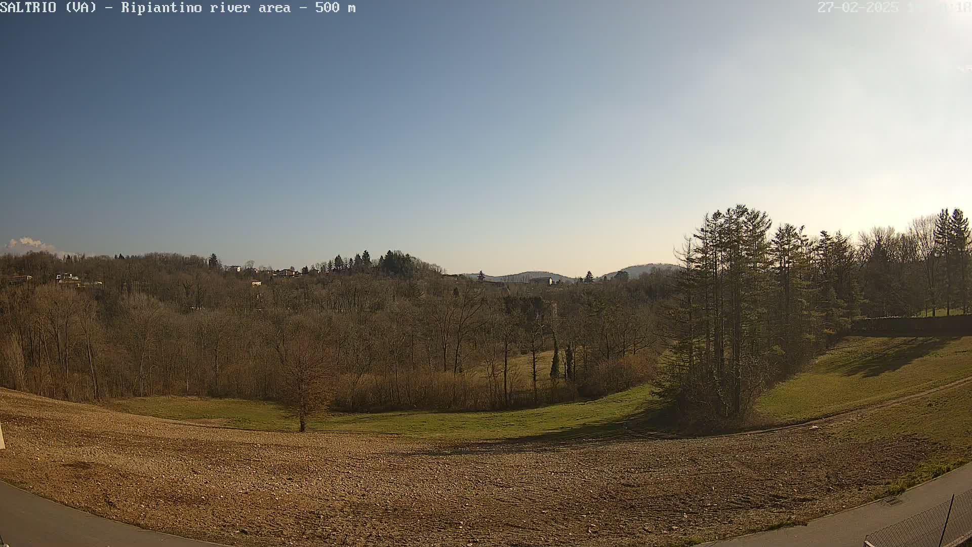 A mostly clear, sunny day overlooks a plowed field, barren trees, and a grassy area leading to a wooded hillside.
