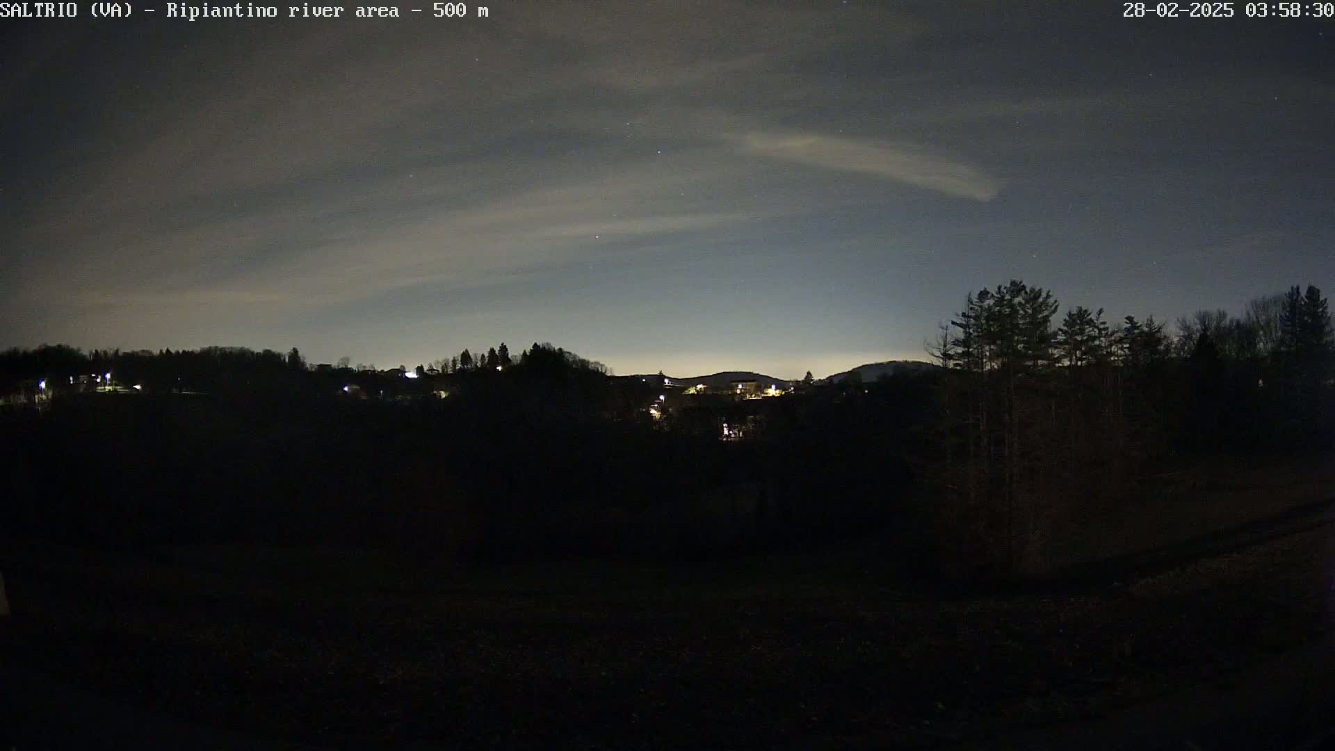 A nighttime view of a dark, sparsely lit landscape with trees and a few distant houses under a mostly clear sky with faint clouds and stars.