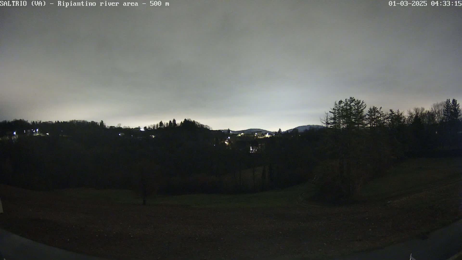 A nighttime view of a dark, tree-covered hillside with a few scattered lights, under a cloudy sky.
